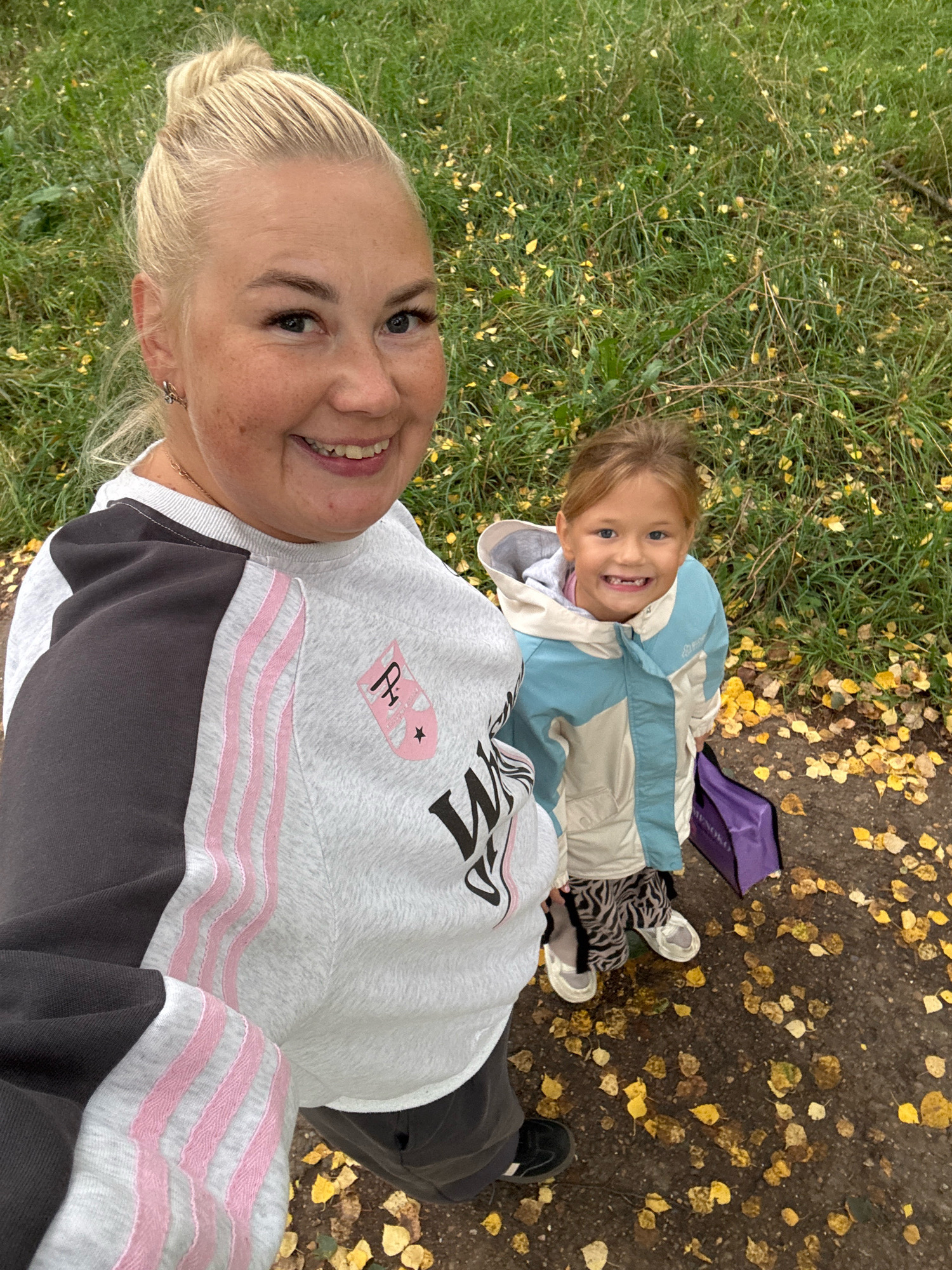 An outdoor, high-angle, selfie-style photo shows two females, an adult and a child, standing on a path covered with fallen yellow leaves. The adult, on the left, has blonde hair pulled up in a bun and is wearing a grey and white sweatshirt with pink stripes. The child, on the right, is wearing a blue and white jacket and black and white patterned pants. The background consists of green grass. The photo appears to have been taken in Chaikovsky, Russia, as indicated by the provided location information.