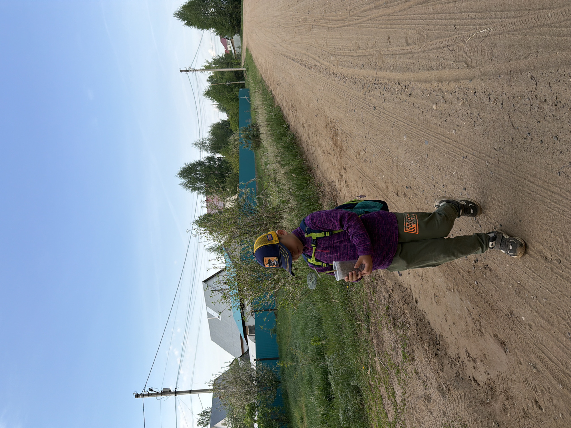 A child is standing on a dirt road in a rural area. The child wears a purple shirt, green pants with "SPORT" on the leg, and a yellow and blue baseball cap. They are carrying a clear plastic container. A green backpack is on their back. The road is sandy and dusty, with tire tracks visible. A green fence lines one side of the road. Houses with blue and gray exteriors are visible behind the fence. A utility pole is visible in the distance. The sky is blue. Based on the background, this scene likely takes place in a rural area near Chaikovsky, Russia.