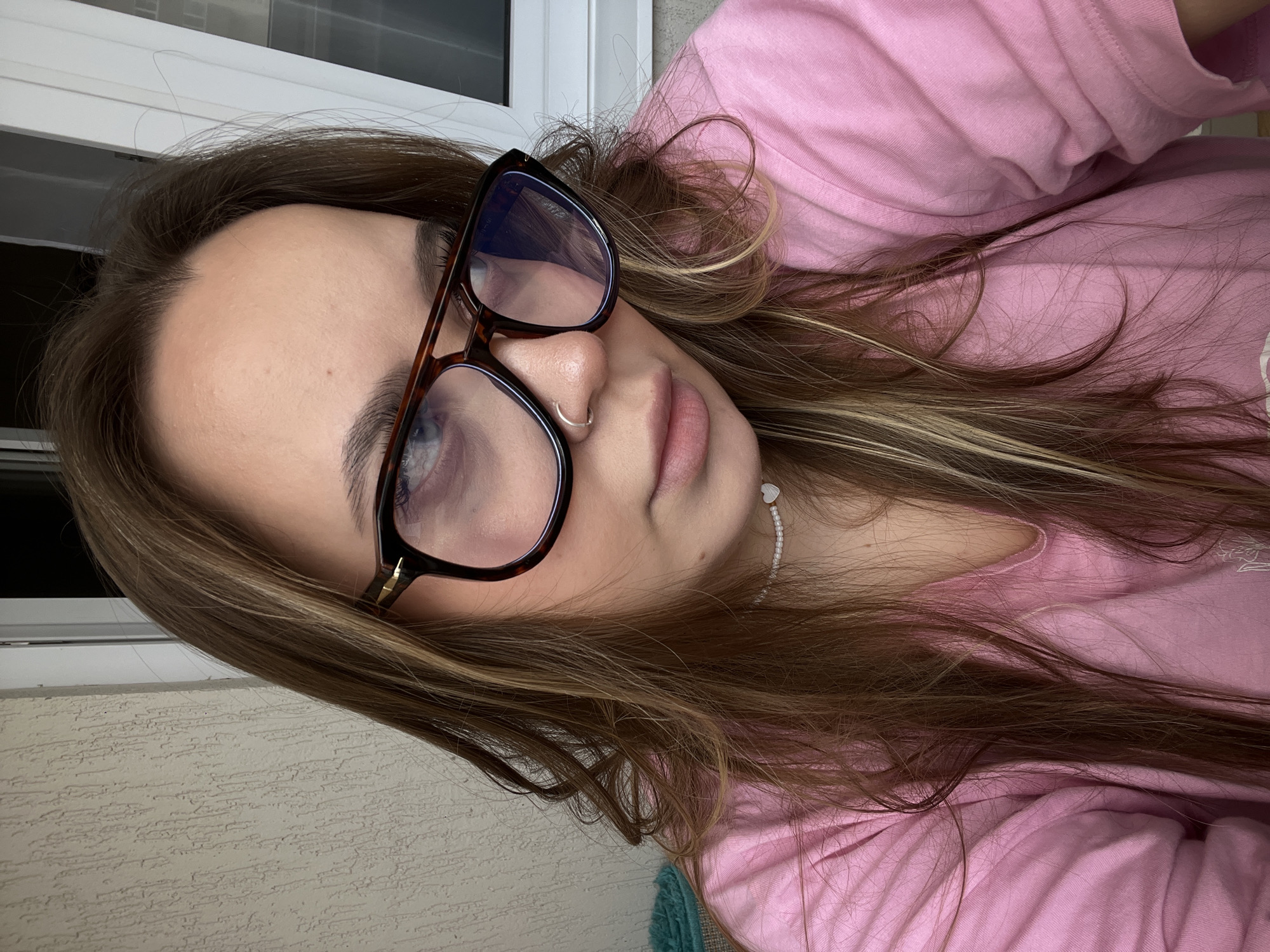 A woman with brown hair, wearing eyeglasses and a pink shirt, is visible in the image. She has a nose ring and is wearing a white beaded necklace. The background shows a textured wall and a window frame. The location is potentially Prigorodny, Russia.