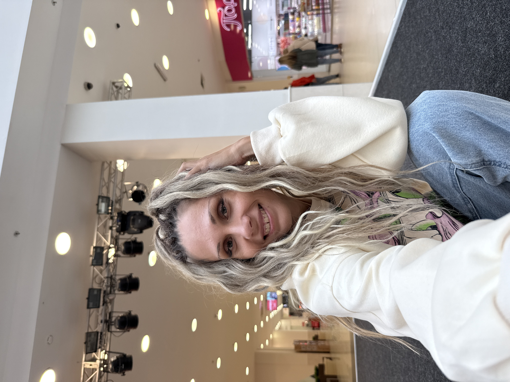 In Rusanovo, Russia, a woman with blonde hair styled in braids smiles as she takes a selfie in an indoor shopping mall. She is wearing a cream-colored sweatshirt and blue jeans. The background shows a brightly lit mall interior with shops and other shoppers.