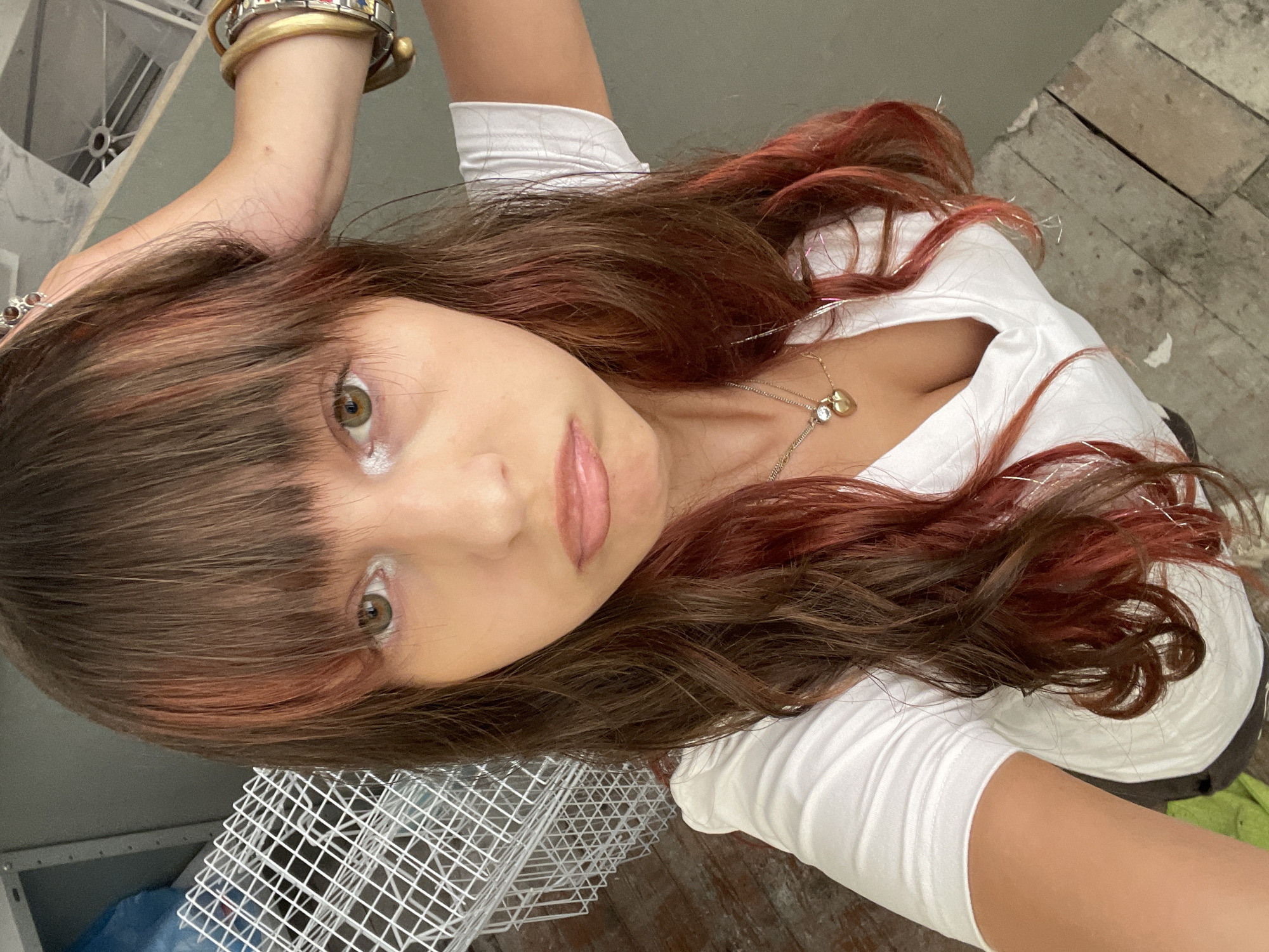 The image is a close-up selfie of a young woman with long, wavy brown hair with reddish highlights. She is wearing a white V-neck t-shirt and several necklaces, one of which has a heart-shaped pendant. Her left hand is raised and resting on her head, and she is wearing multiple bracelets on her right wrist. Her expression is somewhat sullen, with her lips pursed. The background is somewhat blurred, showing a white wire rack and a gray wall. The floor appears to be made of wooden planks. The image does not contain any information to determine its location in Raanana, Israel.