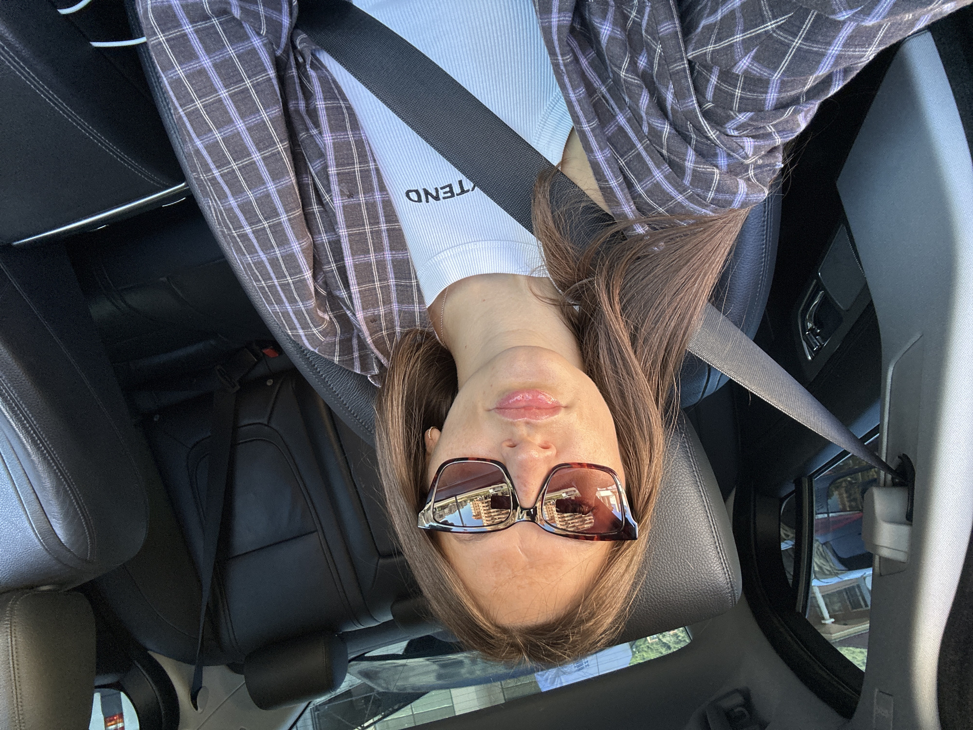 A woman is sitting in the passenger seat of a car, wearing sunglasses, a plaid jacket, and a white ribbed top. The interior of the car is black leather. The woman is looking at the camera, with her mouth slightly open. The car is parked on a street in Donji Pobori, Montenegro, with buildings visible in the background.