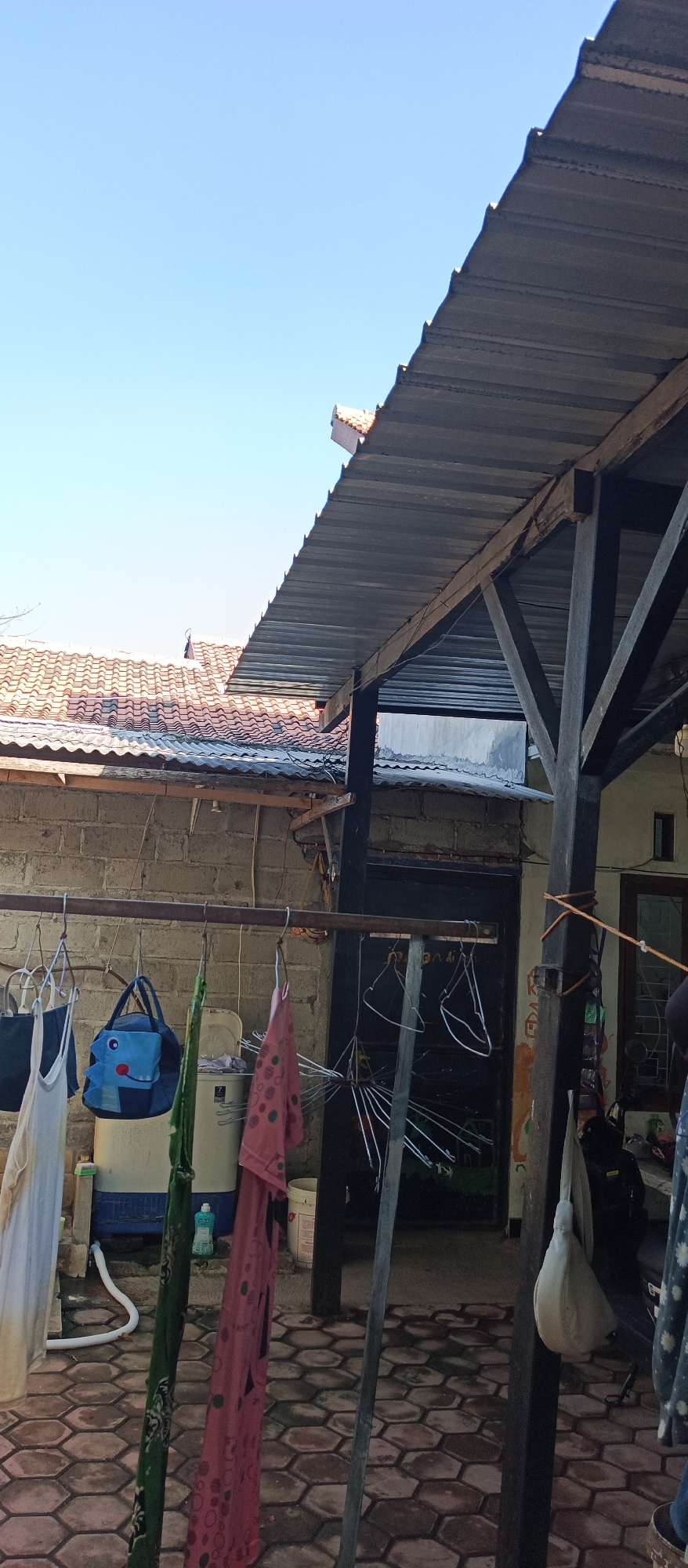 An outdoor scene in Jatisari, Indonesia, features a clothesline laden with various garments, including a white tank top, a blue bag with a cartoon face, a green patterned fabric, and a pink patterned dress. These items are hanging from a metal rod supported by a brick wall. To the right, a part of a corrugated metal roof is visible, supported by dark wooden beams. In the background, a washing machine is partially visible, along with a black door and a window. The ground is paved with hexagonal tiles. The sky above is clear and blue.