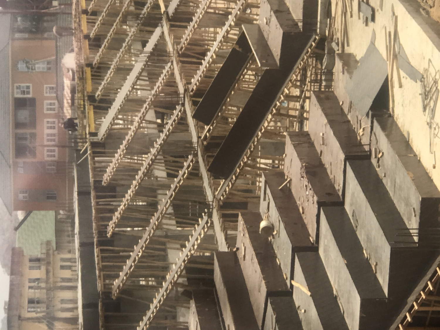 The image displays an active construction site, viewed from an elevated perspective looking down. The central focus is a complex framework of wooden scaffolding and dark concrete forms, which appear to be prepared for pouring, forming the structure for new floors or beams of a building. Wooden support beams crisscross the scaffolding.
One construction worker, dressed in dark clothing, is clearly visible on the scaffolding in the mid-left section, seemingly engaged in building activities. Another less distinct figure might be present further into the site. A small, light-colored cylindrical object, possibly a bucket, hangs from one of the concrete forms on the right side.
In the background, to the left of the construction, several multi-story buildings with numerous windows are visible. These established structures, with facades in shades of brown, orange, and beige, suggest an urban or suburban setting.
The scene is brightly lit by direct sunlight, casting clear shadows and indicating a clear, sunny daytime. The image itself is rotated 90 degrees counter-clockwise. There is no visible text in the image.