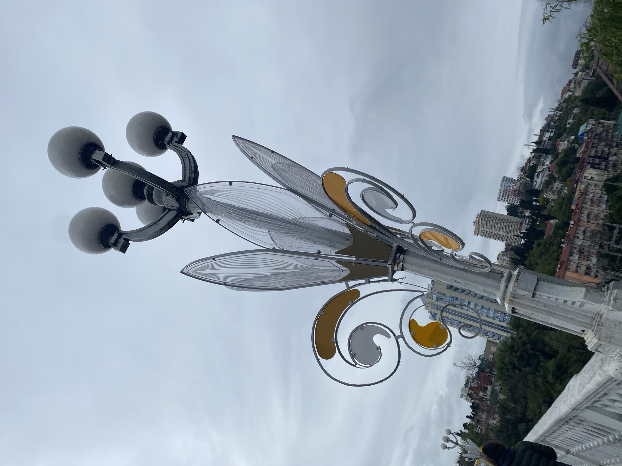 The image depicts a decorative street lamp in Sochi, Russia. The lamp consists of a cluster of six globe-shaped lights attached to a pole adorned with stylized metalwork resembling leaves and swirls with yellow and gray accents. The pole is white and has a decorative, classical design.
In the background, the cityscape of Sochi is visible. Buildings of varying heights and architectural styles can be seen interspersed with trees and vegetation, indicating a hillside or elevated terrain. The sky is overcast with a gray, cloudy expanse. The photograph is taken from a low angle, looking upward at the lamp and cityscape. A portion of a bridge is visible in the lower part of the image.