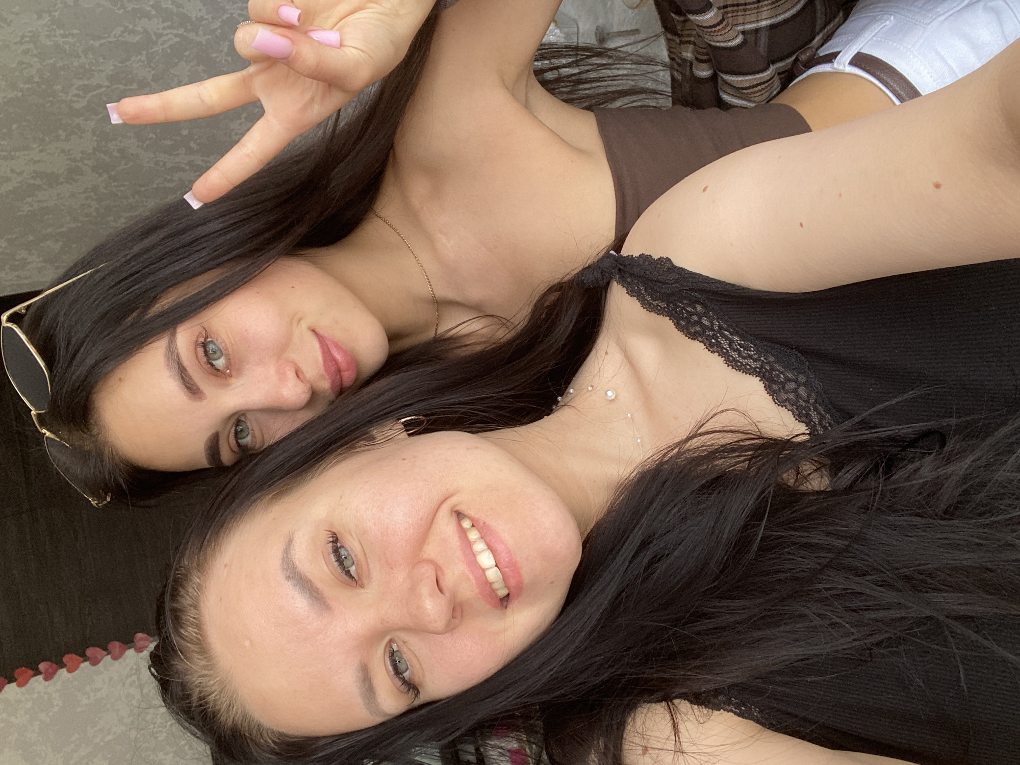 Two young women are posing for a selfie. One woman is wearing a brown top and holding up a peace sign. The other woman is wearing a black tank top with lace trim. The location is unknown and there is no information indicating this image was taken in Chaikovsky, Russia.