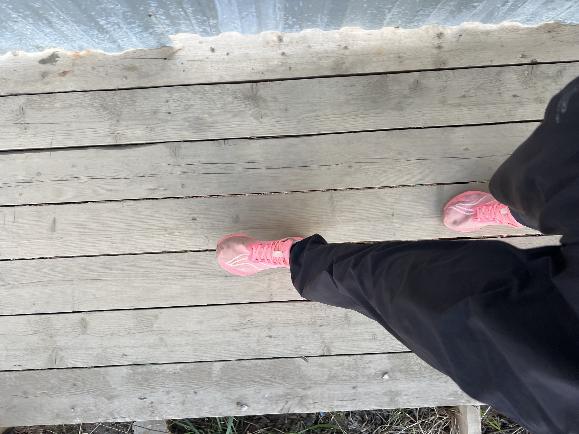 The image shows a person standing on a weathered wooden deck. The deck is composed of horizontal planks with visible grain and occasional knots, painted a light gray color. The person is wearing black pants and pink athletic shoes. The top of the image shows the bottom edge of a corrugated plastic or metal sheet, possibly a roof overhang. The bottom edge of the image shows grass and debris, suggesting an outdoor setting. The location is not evident from the provided visual information and cannot be identified as Perm, Russia without additional context.
