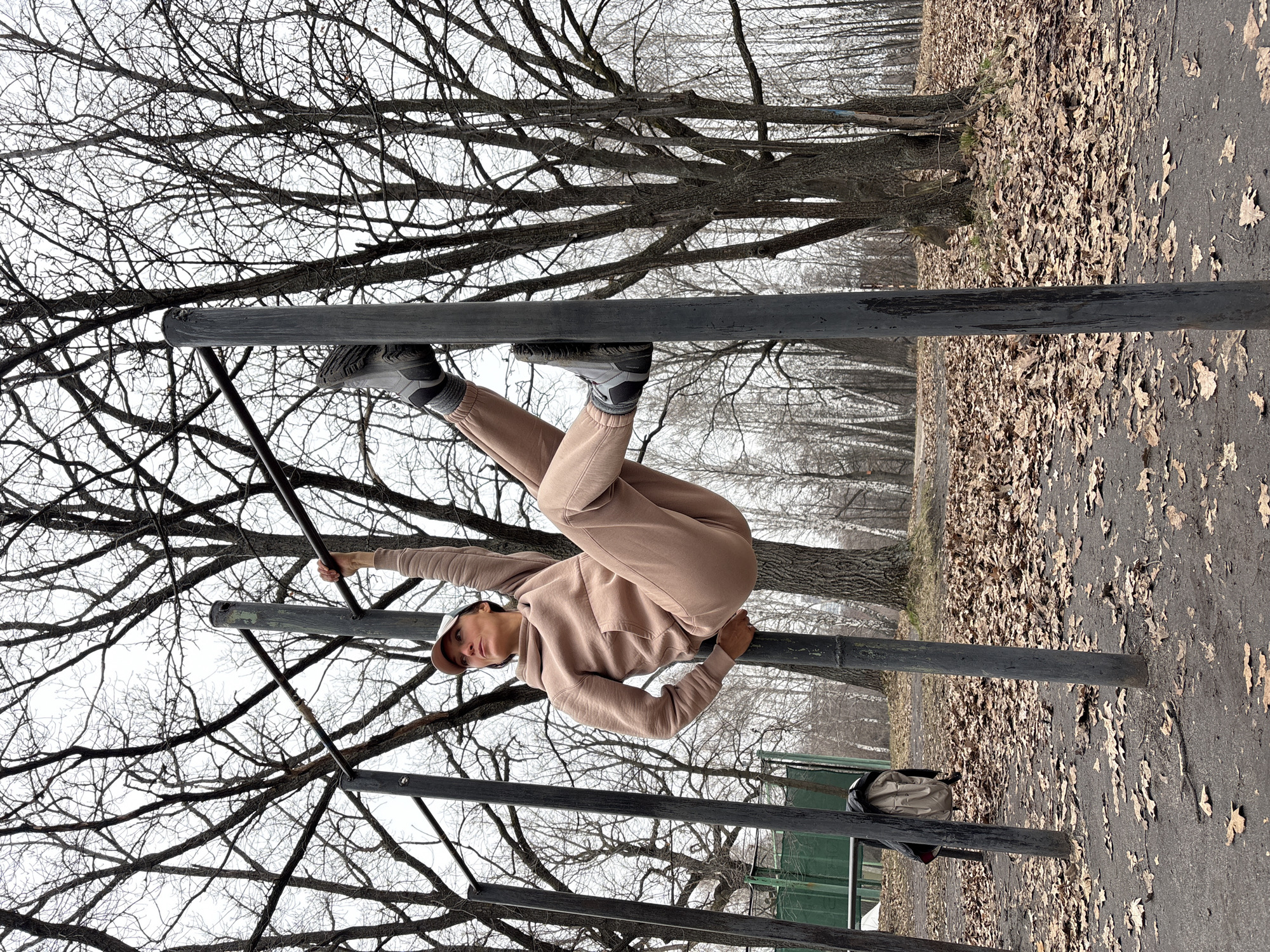 A person is performing a calisthenics exercise on parallel bars in a park in Ulyanovsk, Russia. The person, wearing a beige tracksuit, grey trainers and a cap, has their legs wrapped around one bar and is holding onto another bar with one hand. The background consists of leafless trees and a ground covered with fallen leaves. A tan colored bag is attached to the bar behind them.