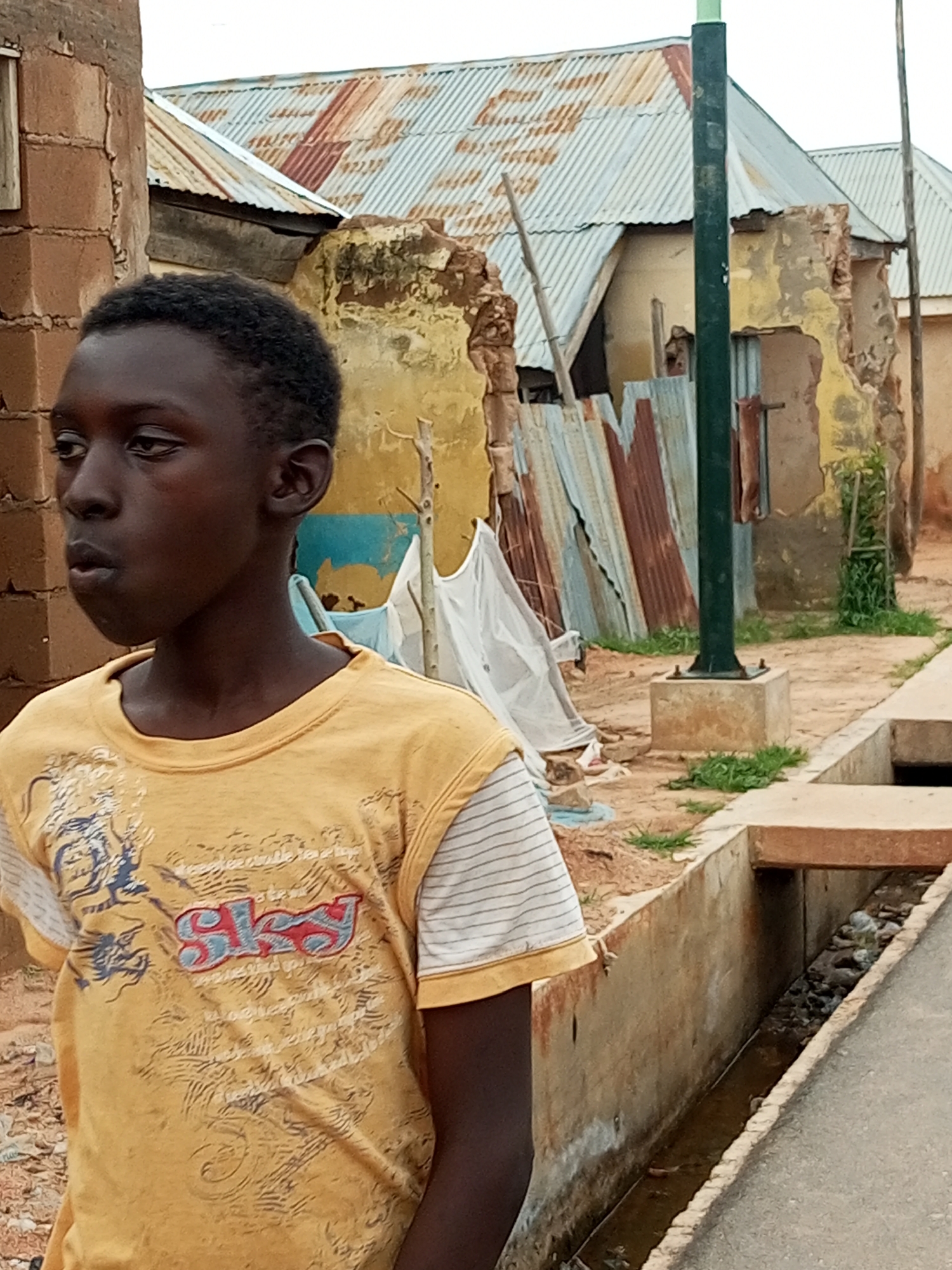 A medium shot captures a young person standing outdoors in Gombe, Nigeria. The individual, with dark skin and short black hair, is wearing a yellow t-shirt with a white striped sleeve and a graphic design on the front. The background depicts a street scene characterized by weathered buildings with corrugated metal roofs, some showing signs of disrepair with exposed brickwork and peeling paint. A dark green pole stands prominently in the middle ground, possibly a lamppost, mounted on a concrete base. A concrete drain runs along the right side of the image, containing murky water and debris. The ground is a mixture of dirt and sparse vegetation. The overall impression is one of a typical urban environment in Gombe, with elements of both everyday life and perhaps some infrastructural challenges.