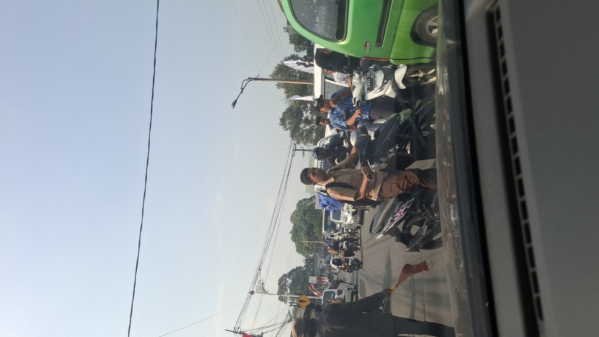 The image depicts a traffic jam on a sunny day, likely in Cilaku, Indonesia, based on the presence of various vehicles and the general urban street scene. In the foreground, a driver's view from inside a vehicle is visible, with the dashboard and windshield dominating the bottom of the frame.
The main subject of the image is the busy street, filled with a variety of vehicles, predominantly motorcycles. Several individuals are riding motorcycles, some appearing to be students in uniforms. One young person in a tan uniform is prominently featured, riding a black motorcycle. Other motorcyclists are also visible, some with passengers. Behind the motorcycles, there are cars and what appears to be a green van on the right side of the frame. Traffic signals and power lines crisscross the sky, indicating an urban environment.
On the left side of the road, a person in dark clothing, possibly a traffic controller, is holding a red flag, suggesting an organized event or a deliberate traffic management situation. Several flags are also visible on poles along the roadside, some white and some with red and white elements, potentially indicating a local celebration or a political event. Trees and buildings can be seen in the background, further confirming the urban setting. The overall atmosphere is one of congestion and activity typical of a busy city.