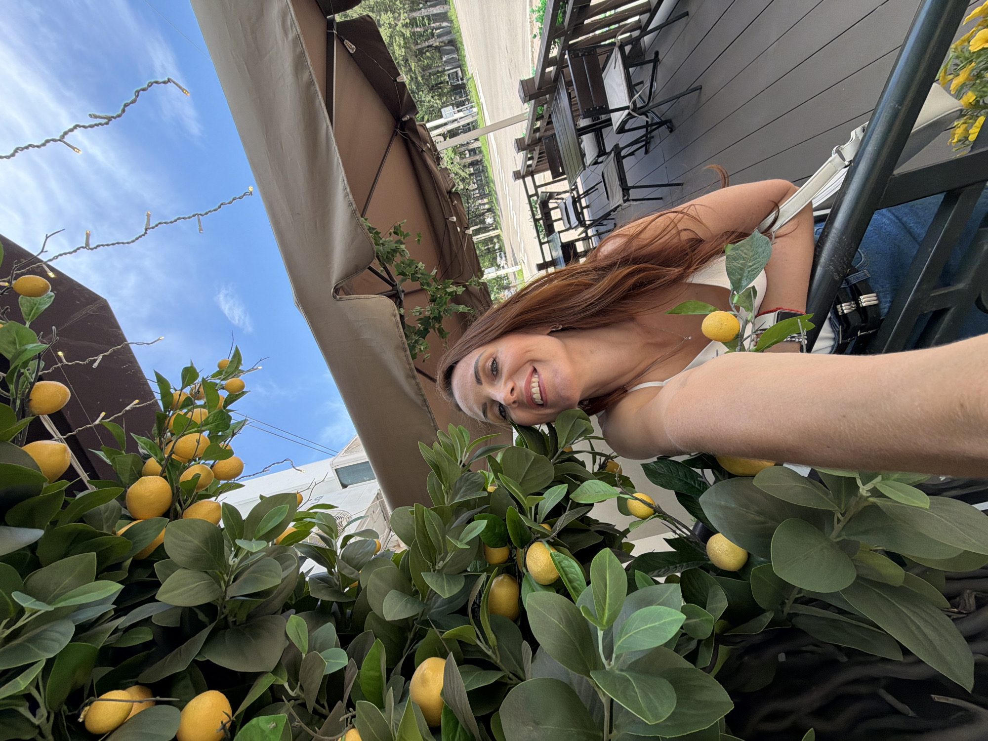A woman with long auburn hair and a white top poses for a selfie in front of a fake lemon tree. She is smiling at the camera, holding a small branch with a lemon and leaves. Behind her, an outdoor seating area with tables and chairs is visible under a large umbrella. In the background, a road with trees and buildings suggests an urban or suburban setting. The sky above is blue with some clouds, and string lights are draped across the scene. The image appears to be taken during the daytime. There is no indication of the location being in Birobidzhan, Russia.