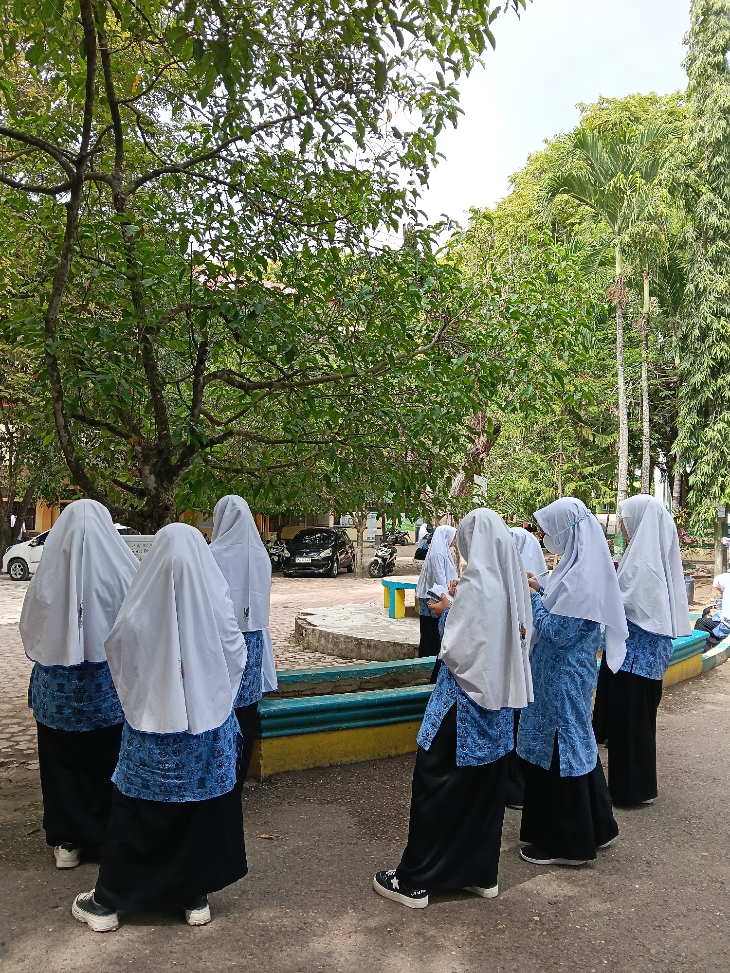 A group of seven female students, all wearing white hijabs and blue patterned blouses with black skirts, are standing outside under a large, leafy tree. Some of the students are holding books or phones. In the background, there is a paved area with cars, a motorcycle, and a park bench. The setting appears to be a schoolyard in Lhokseumawe, Indonesia, given the presence of students in uniforms typical of Indonesian schools and the surrounding tropical vegetation.