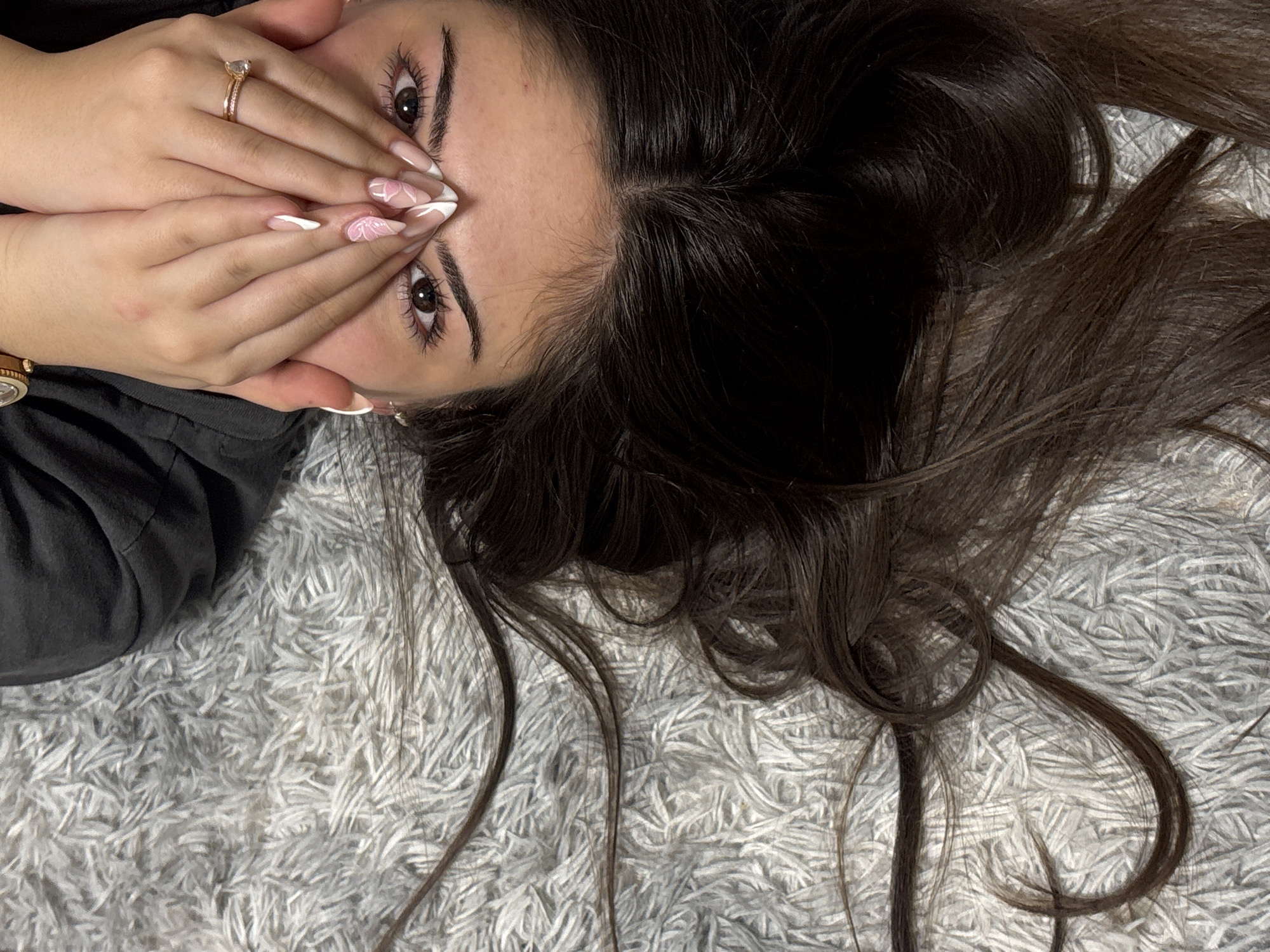 The image depicts a person with long, dark brown hair lying on a white, fluffy rug. The person's face is partially obscured by their hands, which are adorned with a ring and manicured nails featuring white and pink designs. Only the person's eyes and forehead are visible. The person is wearing a dark-colored top. The location is unknown and there is no indication this image was taken in Lehchevo, Bulgaria.