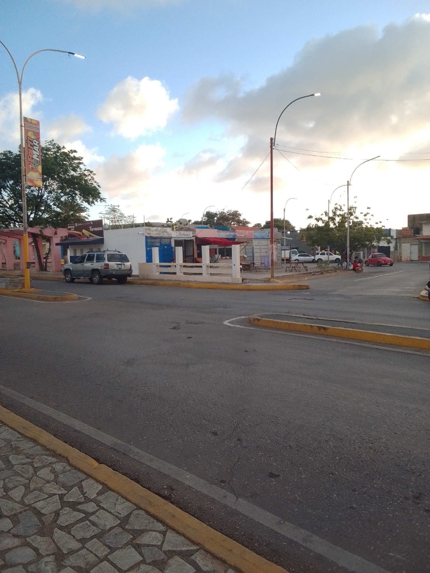 The image depicts a street scene in La Tagua, Venezuela, likely during the late afternoon or early evening, given the lighting. In the foreground, a paved road with yellow painted curbs dominates the lower half of the frame. To the left, a sidewalk paved with irregular stone tiles is visible.
In the mid-ground, a silver SUV is parked on the side of the road. Behind the SUV, a row of low-rise buildings lines the street. These buildings appear to be commercial establishments, with one displaying a sign that reads "Farmacia" (pharmacy). Further down the street, there are more buildings, some with awnings, and a red Volkswagen Beetle is parked. Several streetlights are visible throughout the scene. The sky above is a mix of blue and white clouds, with a hint of the setting sun. The environment suggests a moderately developed urban or semi-urban area.
