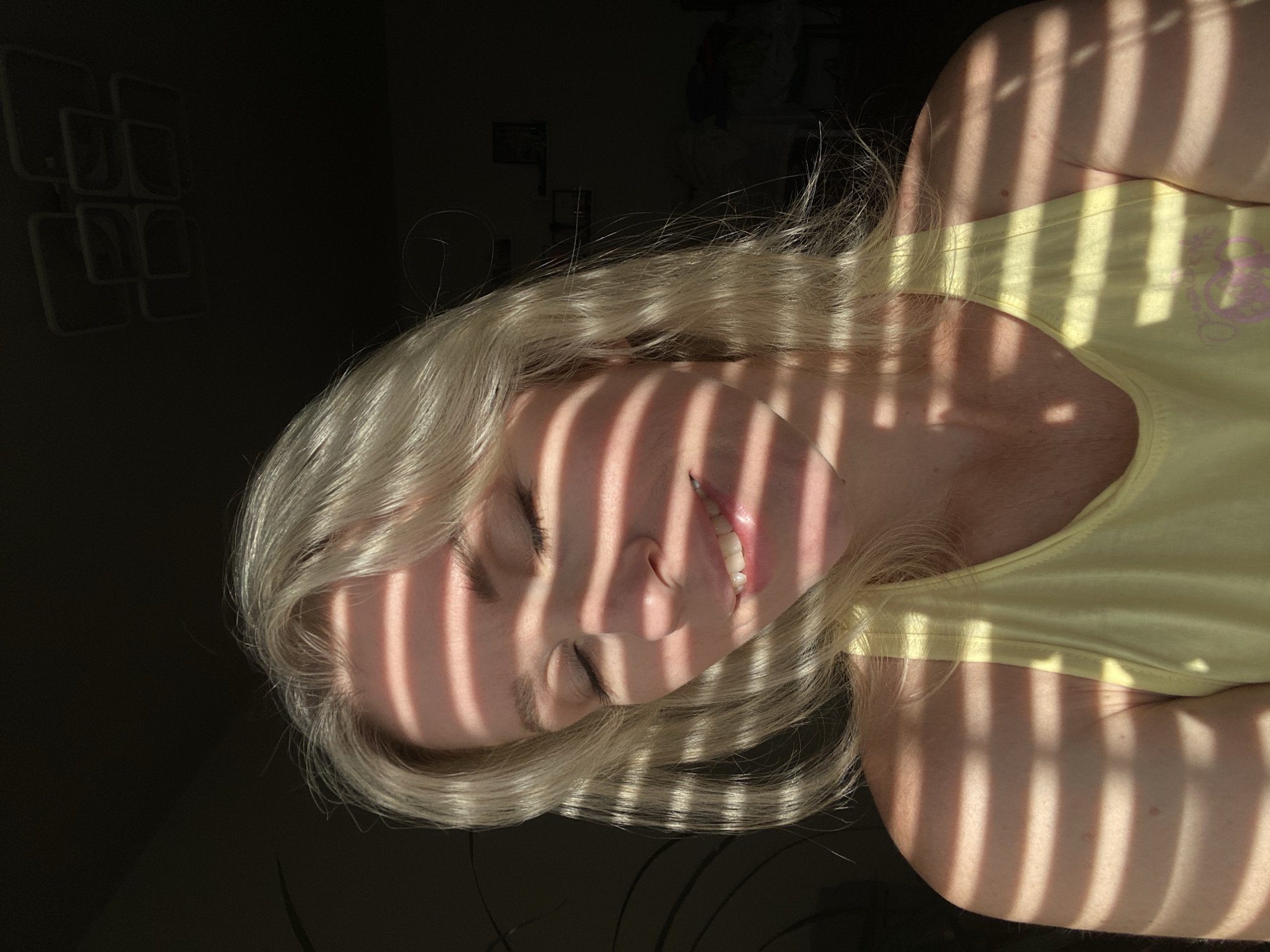 A woman with blonde hair is seen in a close-up, shoulder-up shot. She is wearing a yellow tank top and has her eyes closed, with a gentle smile on her face. Sunlight filters through blinds, casting horizontal stripes of light and shadow across her face and upper body. The background is dark and out of focus, with some indistinct shapes visible. The lighting suggests it is daytime, possibly indoors with sunlight coming through a window.