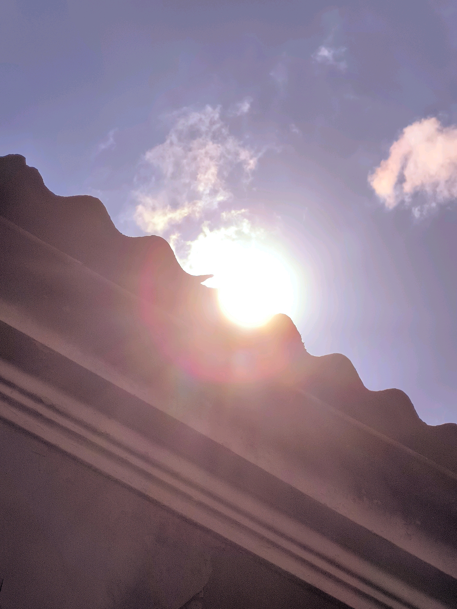The image shows a low-angle view of a corrugated roof against a sky with scattered clouds and the sun. The sun is bright and located to the right of the frame, partially obscured by the roofline and creating lens flare. The sky is a pale blue with some purplish hues, suggesting it might be sunrise or sunset. There are some pinkish clouds on the right side. The roof is a dark, reddish-brown color with distinct horizontal ridges and a jagged edge where it meets the sky. The scene appears to be an outdoor urban environment, possibly in Batumi, Georgia, given the architectural style of the roof, which is common in older residential areas of the city. There are no people or animals visible in the frame. The image captures a moment of natural light interacting with man-made structures.