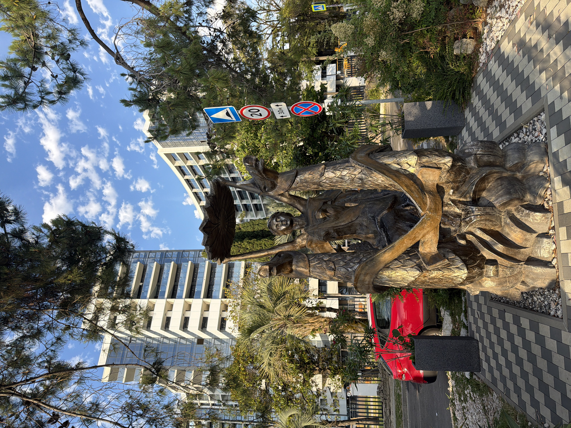 This is an outdoor, eye-level, medium shot of a bronze sculpture in Sochi, Russia. The sculpture depicts a central human figure holding up a circular object above its head, with two dragon-like creatures flanking it. The sculpture is located on a paved pathway with checkerboard-patterned tiles, surrounded by gravel and vegetation, including pine trees and palm trees. In the background, there are modern, multi-story buildings with balconies and glass facades, as well as a red car parked on the street. Several traffic signs are visible on a pole next to the sculpture, including a pedestrian crossing sign, a speed limit sign indicating 20, and a no stopping sign. The sky is blue with scattered white clouds. The scene appears to be a public area, possibly a park or a promenade in a resort town.