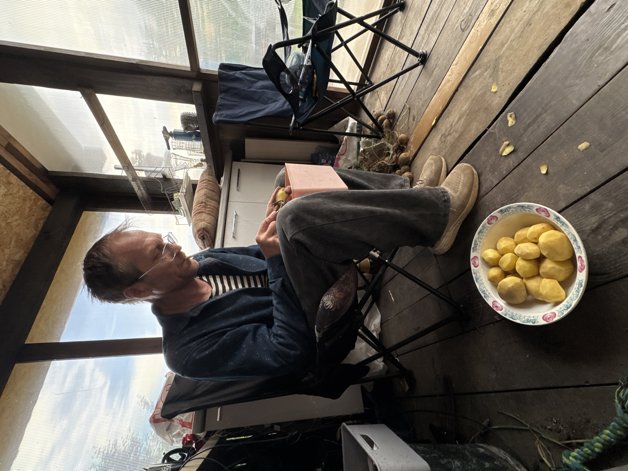 The image captures a man seated in a portable folding chair on a wooden deck inside an enclosed space, possibly a greenhouse or porch-like structure in Chaikovsky, Russia. The man is wearing a striped shirt, a blue jacket, grey pants, and beige shoes. He is holding a small yellow object, likely a potato, in his hands and appears to be peeling it with a tool that is not clearly visible. A pink tablet-like object rests on his lap.
Next to him, on the wooden deck, is a bowl filled with peeled potatoes. Nearby, there are unpeeled potatoes scattered on the deck floor. In the background, there's another folding chair and what appears to be a white cabinet or appliance against the wall. The enclosed space has a translucent roof and walls, allowing natural light to illuminate the scene. The exterior view through the translucent panels suggests an outdoor environment.
