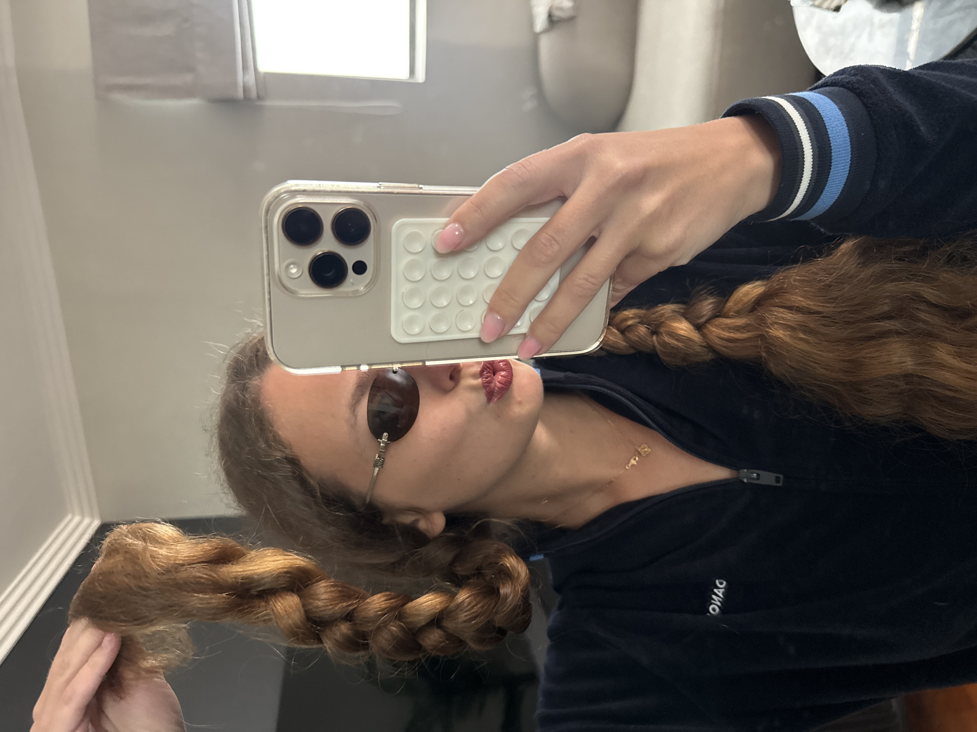 A woman with long braided hair is taking a selfie in a bathroom mirror. She is wearing sunglasses and a dark blue zip-up jacket with light blue and white stripes on the sleeves. The phone she is holding has a light-colored case with a round "pop-it" grip on the back. The background shows a bathroom setting with a window and a toilet. The location in the city of Cape Town, South Africa cannot be determined from the image.