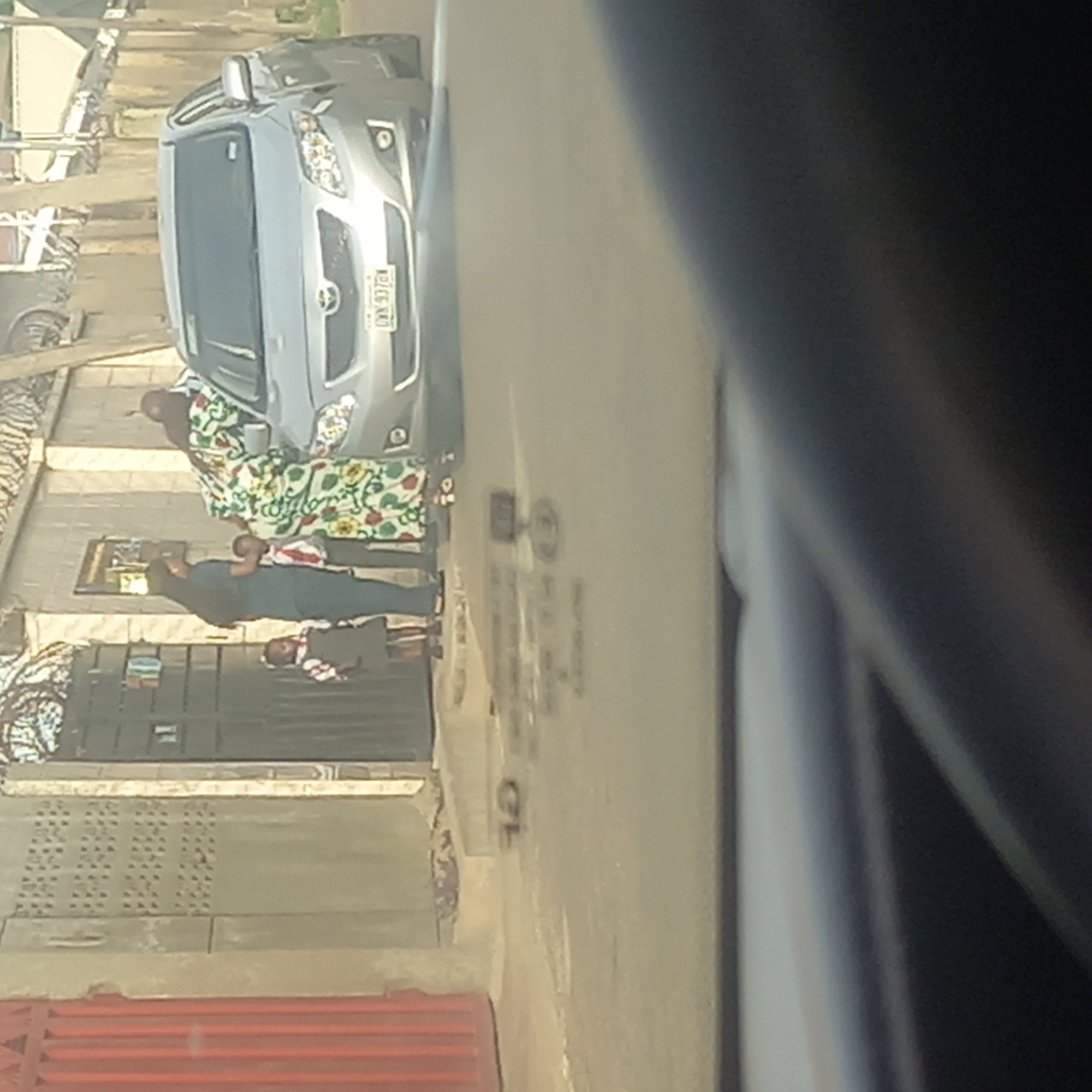 The image depicts a street scene in Barden Yana, Nigeria. A silver car is parked on the side of the road. Adjacent to the car are three individuals: an older woman in a colorful floral patterned dress, a person in dark clothing, and a child in a light-colored top and darker bottoms. The person in dark clothing appears to be carrying the child. The background includes a building with a dark gate and a red corrugated structure at the bottom. The image is captured from inside a vehicle, as evidenced by the interior of a car visible in the foreground.