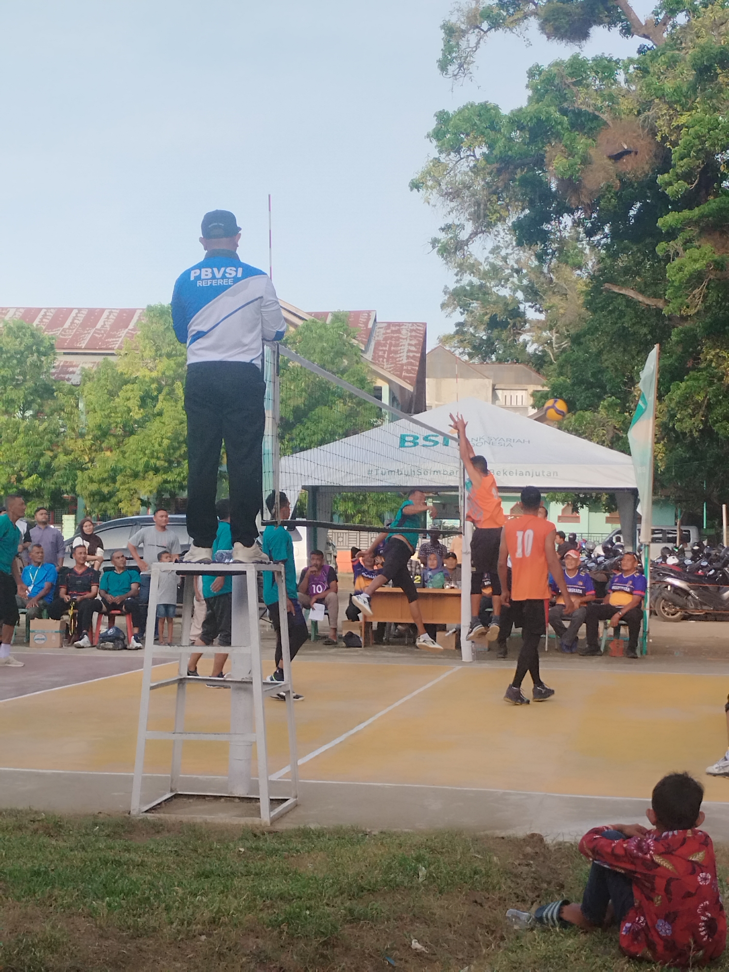 The image depicts a volleyball match in progress, likely in Lhokseumawe, Indonesia, given the presence of the "BANK SYARIAH INDONESIA" tent. The scene is outdoors on a yellow volleyball court. In the foreground, a young boy in a red patterned shirt and dark pants is sitting on the grass, facing away from the camera and towards the game. A referee, wearing a blue and white jacket with "PBVSI REFEREE" clearly visible, stands elevated on a white platform to the left, observing the game.
In the middle ground, players are actively engaged in the match. One player in an orange jersey with the number "10" is positioned on the court, while another player in a green jersey is in mid-air, jumping to hit the ball. The volleyball is visible in the air above the net. Spectators are seated on benches on the left side of the court, watching the game. Behind them, a white tent with the BSI logo is set up. Beyond the court, trees and buildings can be seen. To the far right, a line of motorcycles is parked. The sky is clear and bright.