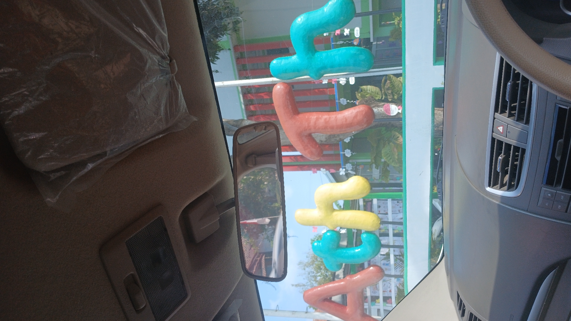 The image shows a view from inside a car, looking out the windshield. The dashboard and steering wheel are visible in the foreground. Through the windshield, there are colorful, puffy letters attached to the glass, spelling out "ACTH". Behind the letters, a building with red and green striped walls is visible, suggesting it could be a school or community center in Gombong, Indonesia. The sky is clear and blue. The overall scene is bright and cheerful, likely taken on a sunny day.