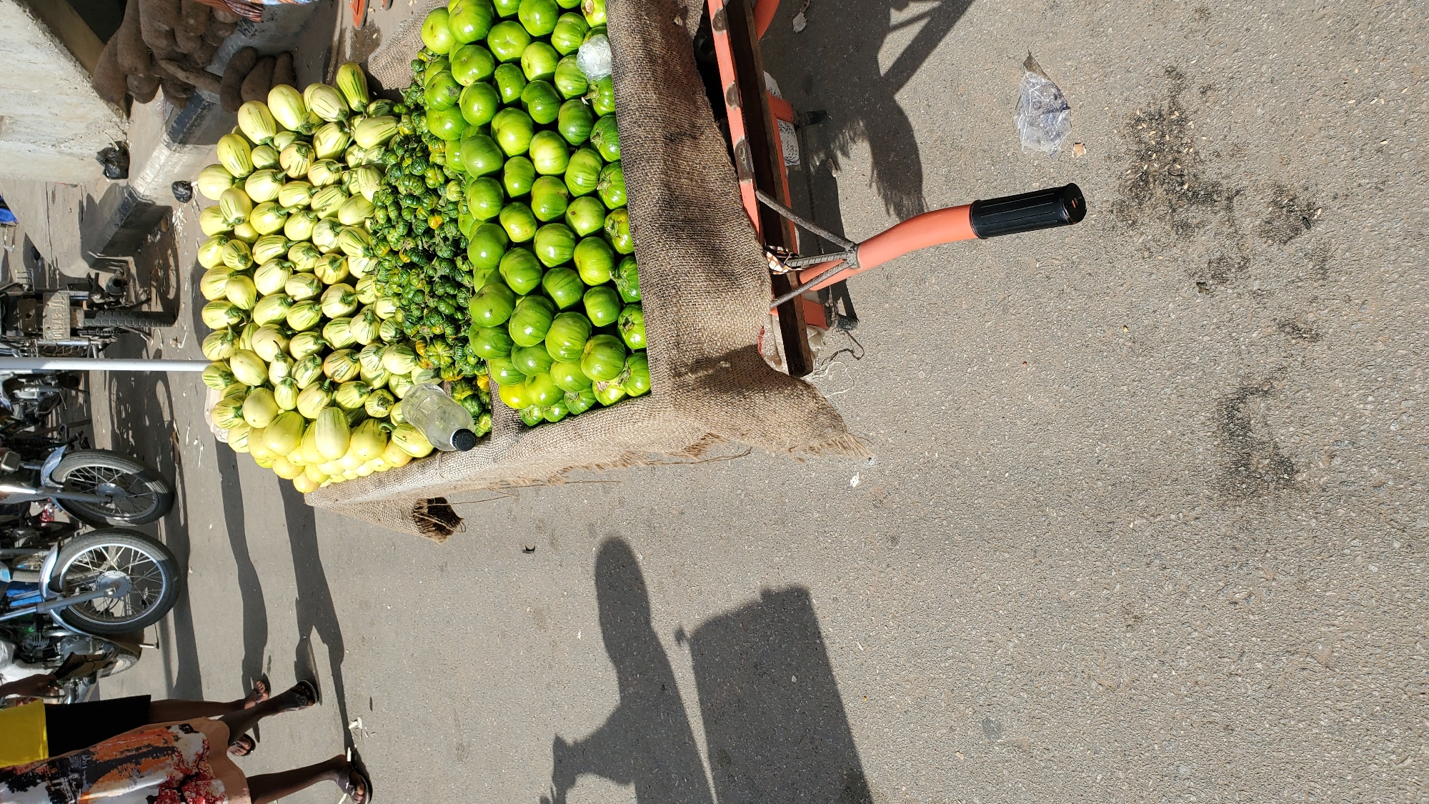 A low-angle, eye-level, full shot captures a street vendor's cart loaded with produce in Jalingo, Nigeria. The cart, made of dark wood and orange metal, is partially covered with burlap. It is filled with a mound of pale yellow and green striped gourds, a smaller pile of small, spiky green gourds, and a large quantity of round, green vegetables, possibly tomatoes or small gourds. A clear plastic bottle lies on its side among the vegetables. In the background, two motorcycles are visible, one with a silver frame and spoked wheels, and another that is dark-colored. A person's legs are visible on the left side of the frame, wearing a patterned skirt and sandals, and another person's legs are partially visible on the right. The cart rests on an asphalt surface, with shadows of people and objects cast across it, indicating a sunny day. The environment appears to be an outdoor market or street scene.