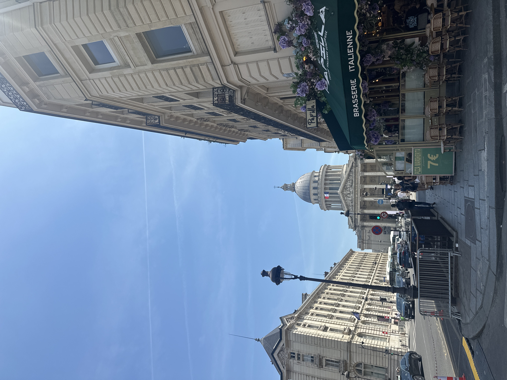 This media file depicts a street scene in Paris, France, on a clear, sunny day. The perspective is looking upwards, showcasing the architecture of Parisian buildings and a glimpse of the sky.
The subjects include several buildings with distinct architectural styles. On the left, a building with light-colored stone facade features multiple rectangular windows and decorative metalwork. In the center, the dome of a prominent building, likely the Panthéon, is visible against the bright blue sky. To the right, another building houses a "BRASSERIE ITALIENNE" with a green awning adorned with purple flowers. Outdoor seating with wicker chairs is arranged in front of the brasserie. Further down the street, more buildings and parked cars line the sidewalk.
The activity appears to be everyday urban life. People are seen walking on the sidewalk and entering or exiting the brasserie. Cars are parked along the street, suggesting ongoing traffic and activity. A street lamp is prominently featured in the foreground of one of the buildings.
Notable details include the bright sunlight indicating it is daytime. The clear blue sky with faint contrails from airplanes suggests good weather. The architectural style of the buildings, the presence of a brasserie with outdoor seating, and the mention of "Paris, France" in the context strongly point to this being a popular tourist area within the city.
Visible text includes "BRASSERIE ITALIENNE" on the awning and "Papila" on a sign beneath the flowers. A sign in front of the brasserie advertises "PIZZA MARGHERITA 7€". Other smaller signs are present but not legible.
The overall impression is one of a vibrant and picturesque Parisian street, likely during a pleasant time of day for outdoor dining and sightseeing.