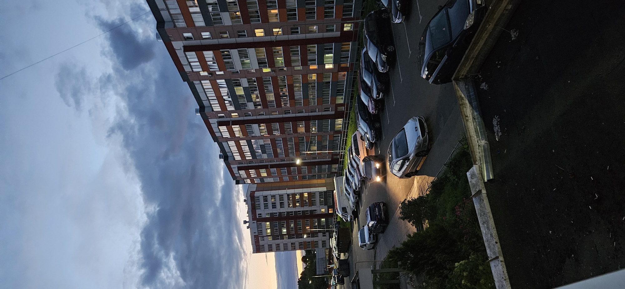 The image depicts a twilight scene in a residential area, likely a courtyard of apartment buildings. Two multi-story apartment buildings with numerous windows, some illuminated, dominate the background. The buildings appear to be constructed with a combination of white, red, and grey facade elements. In the foreground, a paved parking lot is visible, occupied by several parked cars of various makes and models, including a silver Peugeot, a dark SUV, and other vehicles. One car's headlights are on, illuminating the pavement. Adjacent to the parking lot is a strip of greenery and trees. The sky is cloudy with a gradient of colors suggesting the end of daylight. The scene is static and devoid of human activity. Based on the metadata provided, this image was taken in Vitsebsk, Belarus.