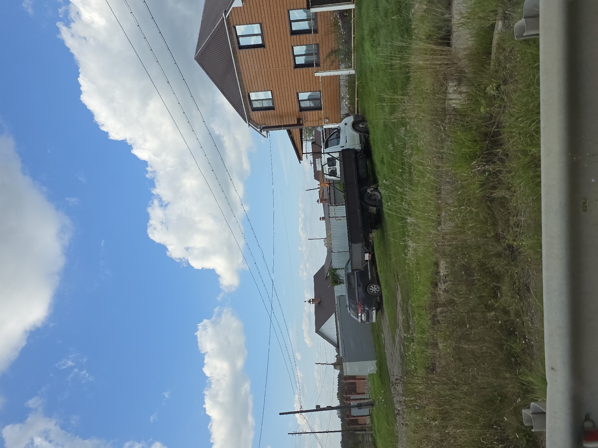 The image captures a suburban or rural scene in Kuluevo, Russia. In the foreground, tall, dry grass and weeds line a dusty dirt road. Behind the road, a dark blue SUV is parked next to a light-colored utility truck with an open bed. The truck is positioned in front of a large, two-story house with brown wooden siding and a dark brown roof. Several windows are visible on the house, reflecting the blue sky and white clouds. To the left of the house, a smaller building with a grey metal exterior is visible, and beyond that, a fence made of red brick and metal posts encloses an area. More houses and fences can be seen in the distance. Overhead, power lines stretch across the frame, connecting to utility poles. The sky is a vibrant blue with fluffy white clouds scattered throughout. The overall impression is a sunny day in a residential area outside of a major city.