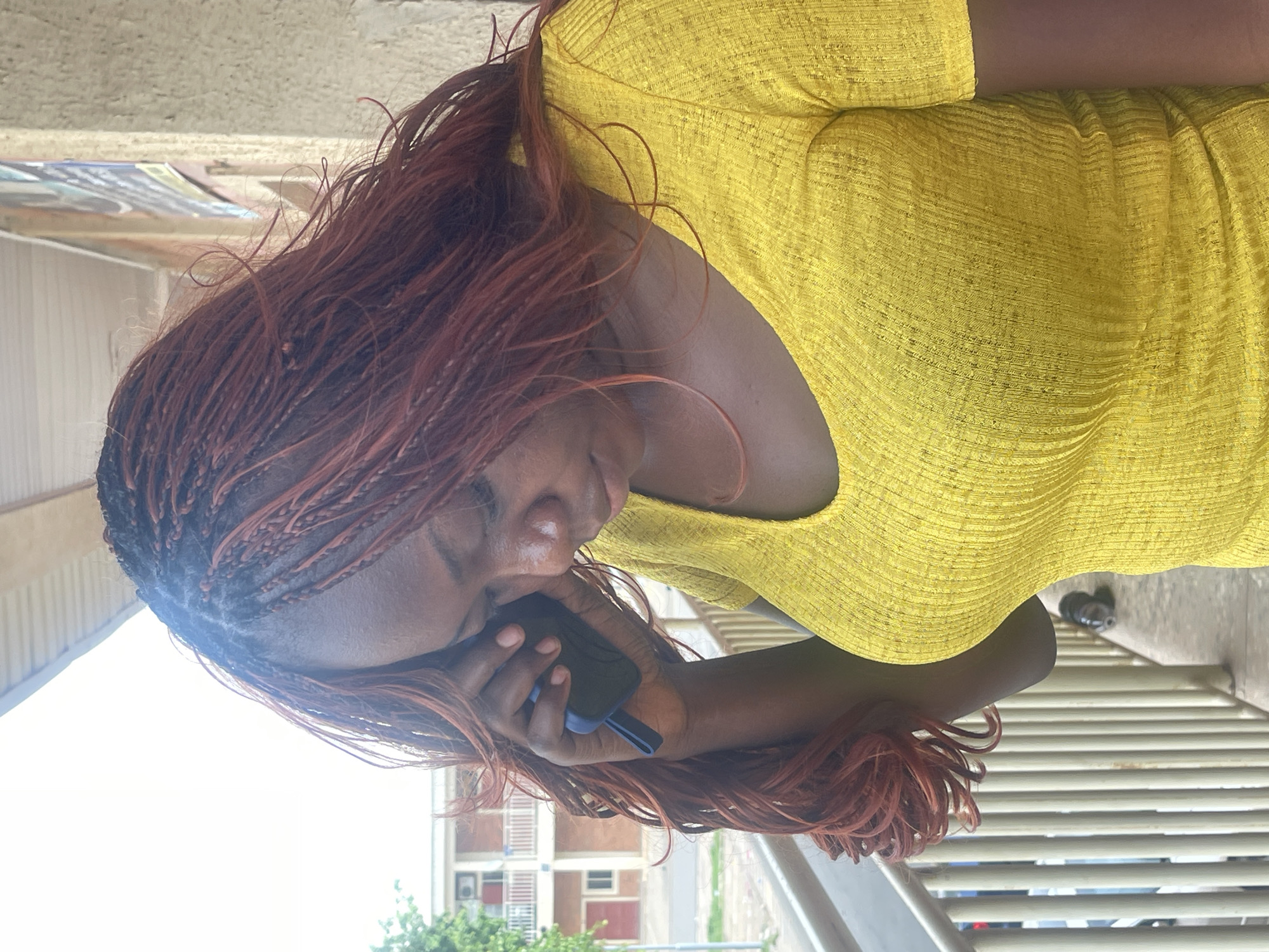 The image, rotated 90 degrees counter-clockwise, captures a young adult with dark skin engaged in a phone call in an outdoor setting, likely in Abuja, Nigeria. The subject is shown in profile, looking downwards with a focused expression.
She has long, reddish-brown hair styled in numerous small, intricate braids. She is wearing a bright, vibrant yellow top or dress, which features a distinct textured, horizontally ribbed fabric. Her left hand holds a dark-colored smartphone to her left ear, with her fingers clearly visible around the device.
The scene is brightly lit by natural daylight, suggesting it is daytime. The background behind her left side is dominated by a bright, almost white sky or light source. To her right, the background includes horizontal railings or slats, indicating an elevated structure such as a balcony, bridge, or walkway. Further in the distance, parts of a building with reddish sections and windows are visible, along with some green trees or bushes.
There is no visible text on the person, clothing, phone, or in the discernable background elements. The image conveys a moment of concentrated communication in an urban outdoor environment.