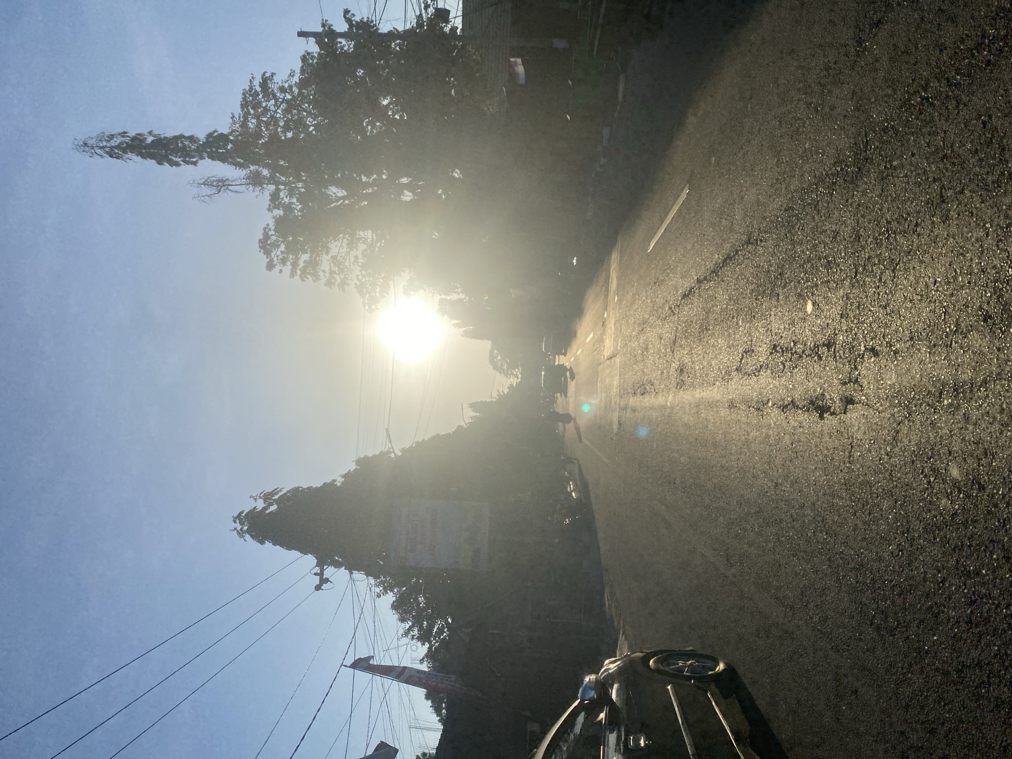The image captures a street scene in Bantaeng, Indonesia, bathed in the bright, hazy glow of the sun during what appears to be sunrise or sunset. The road, paved and with a single white dividing line visible, runs towards the horizon. The sun, positioned slightly left of center in the upper half of the frame, is the dominant light source, creating lens flare and washing out details in its vicinity.
On the left side of the road, a dark-colored car is partially visible, its front end facing away from the viewer. Beyond the car, a flag, appearing red and white, is mounted on a pole, suggesting a national or local celebration. Lined with trees, the street is flanked by buildings and vegetation, with power lines strung overhead. Several trees are prominent, including a particularly tall, slender tree with sparse foliage. Other vehicles, including cars and motorcycles, are present on the road, some moving and others stationary, though they are largely obscured by the glare and distance. In the background, smaller buildings and more trees line the street as it recedes into the distance. The overall impression is that of a typical urban or semi-urban environment in Indonesia, experiencing a bright, atmospheric lighting condition.