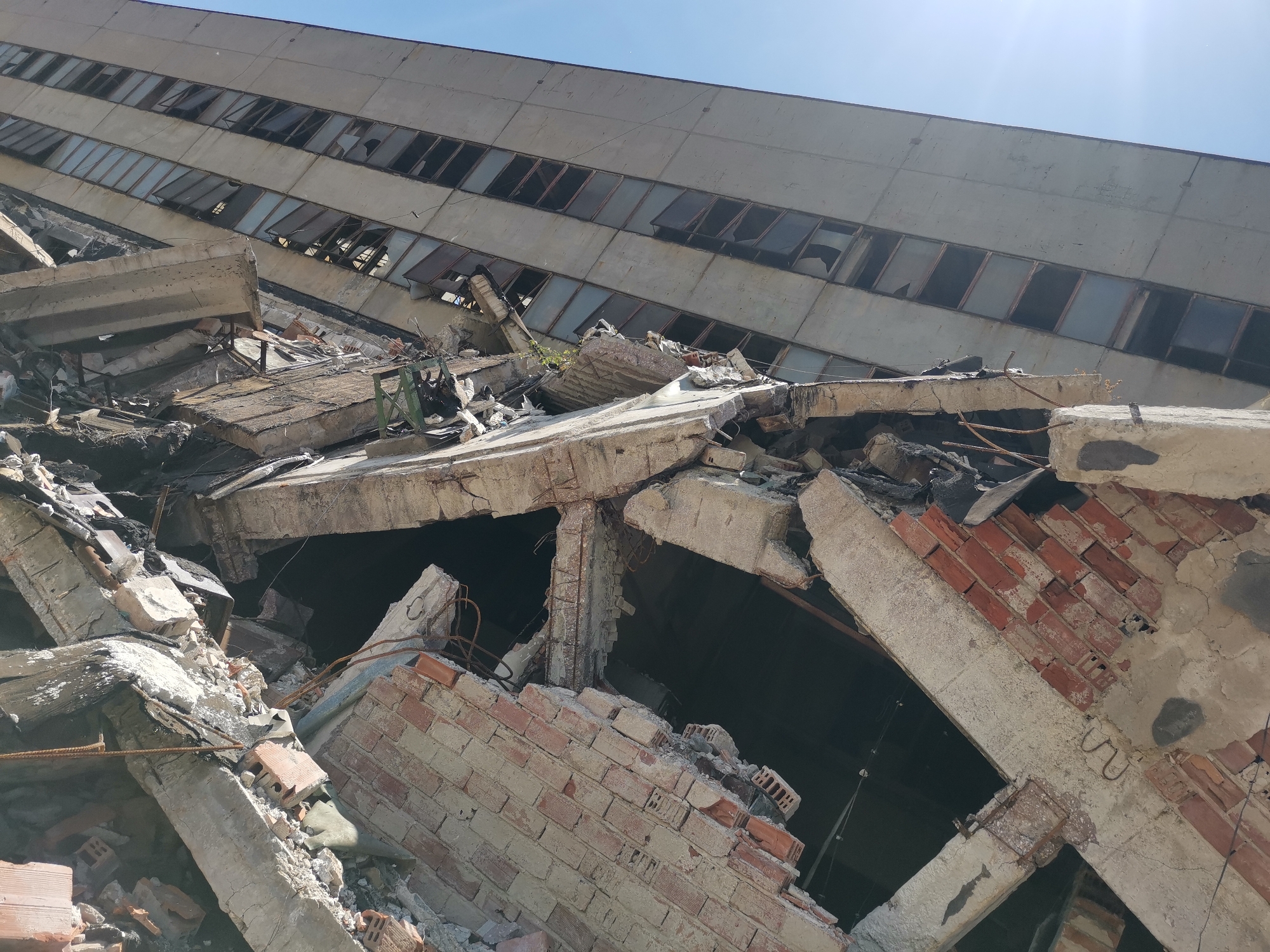 The image depicts the aftermath of severe structural damage to a building, likely caused by a major event such as an earthquake or demolition. In the foreground, a chaotic pile of rubble dominates, composed of broken concrete, twisted rebar, and shattered red bricks. These materials suggest a building constructed with reinforced concrete and brickwork. The rubble obscures any clear view of the ground. In the background, a damaged multi-story building looms. The building has a repetitive pattern of rectangular windows, many of which are broken or missing, revealing the structure's skeleton. The overall color palette is dominated by grays and browns, reflecting the materials of the destroyed buildings. The location of the image is Zhivkovo, Bulgaria. The building and surrounding area are desolate and in ruins. There are no people in the image.