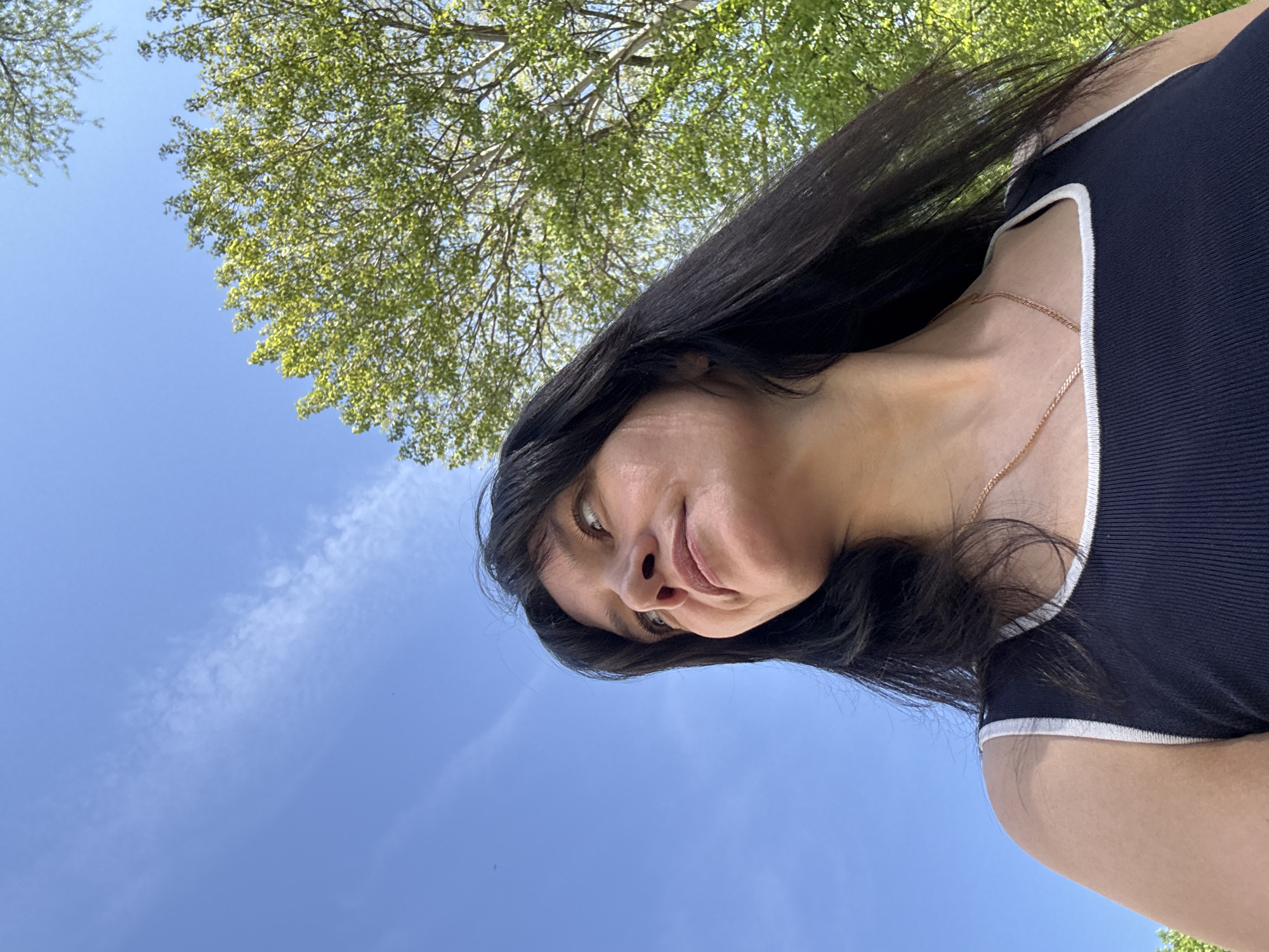 A low-angle shot shows a woman outdoors, likely in a park in Chaikovsky, Russia, as indicated by the prompt. She is facing the camera with a slight smile, her long dark hair falling around her face. She wears a dark top with white trim. A thin gold chain necklace is visible. Above and behind her is a blue sky with wispy clouds and a tree with green leaves. The photo is taken at an angle, tilted approximately 90 degrees clockwise.
