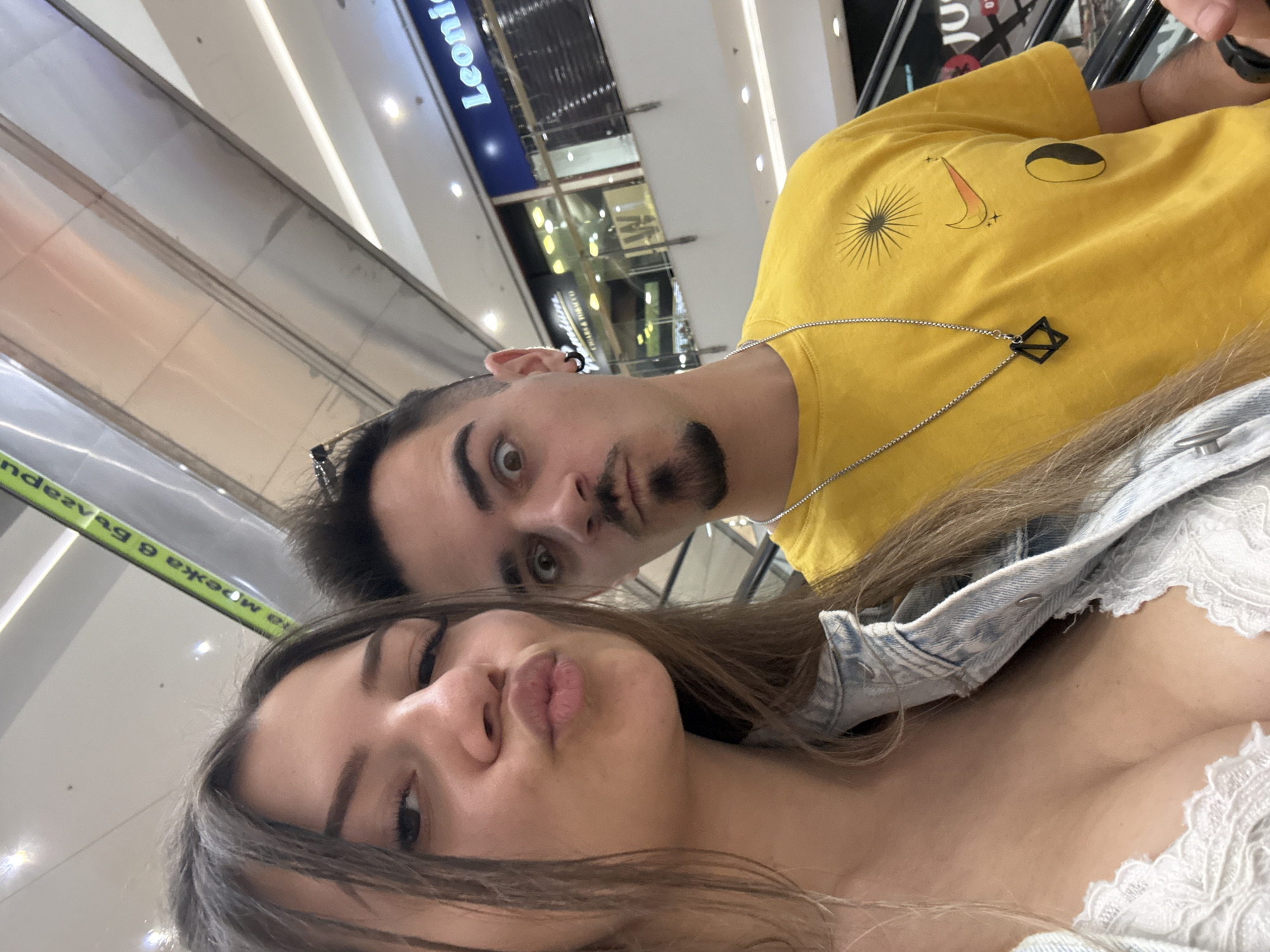 A selfie taken on an escalator in a shopping mall. A young woman with light brown hair is positioned in the foreground, puckering her lips. Behind her, a young man with dark hair and a goatee is looking into the camera with a slightly surprised expression. The man wears a yellow t-shirt with several images. He also wears a black necklace with a square pendant. The woman is wearing a denim jacket and a white lace top. In the background, there is an escalator and signs in Bulgarian, indicating the location is likely Sofia, Bulgaria. The word "Leonid" appears on a blue sign.