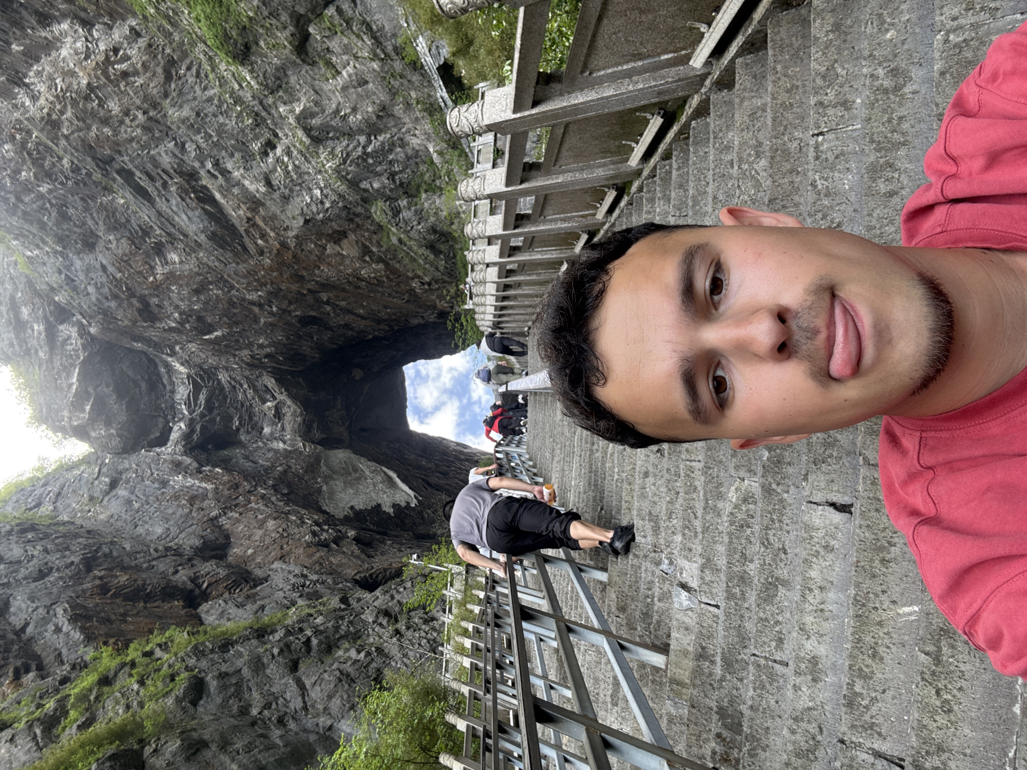 A man takes a selfie at Tianmen Cave, also known as Heaven's Gate, in Zhangjiajie, China. The cave is a large hole in a mountainside. The man is in the upper right corner of the image, wearing a red shirt. He is standing on a gray stone staircase with metal railings, and there are other people on the stairs in front of him. The massive rock formation with the natural arch is to the left. Green vegetation grows on the rocks. The sky is visible through the opening.