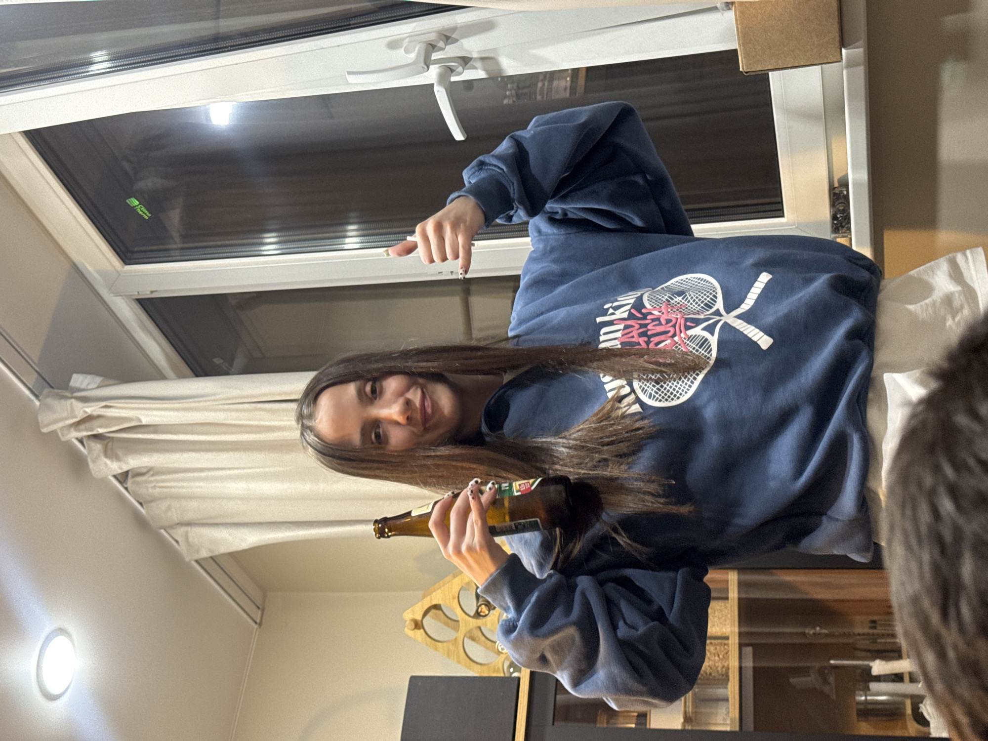 A young woman is indoors, holding a brown glass bottle, possibly beer, in her hands. She is wearing a navy blue sweatshirt with a design of crossed tennis rackets and pink details. She has long brown hair. She is smiling and gesturing towards the window with her left hand. Behind her, there is a window with a white frame and a handle. Curtains are visible to the side. A wooden wine rack is partially visible to the left of her. Behind her, there is a cabinet with items visible inside. The precise location in the city of Razvilka, Russia, cannot be determined from the provided image.