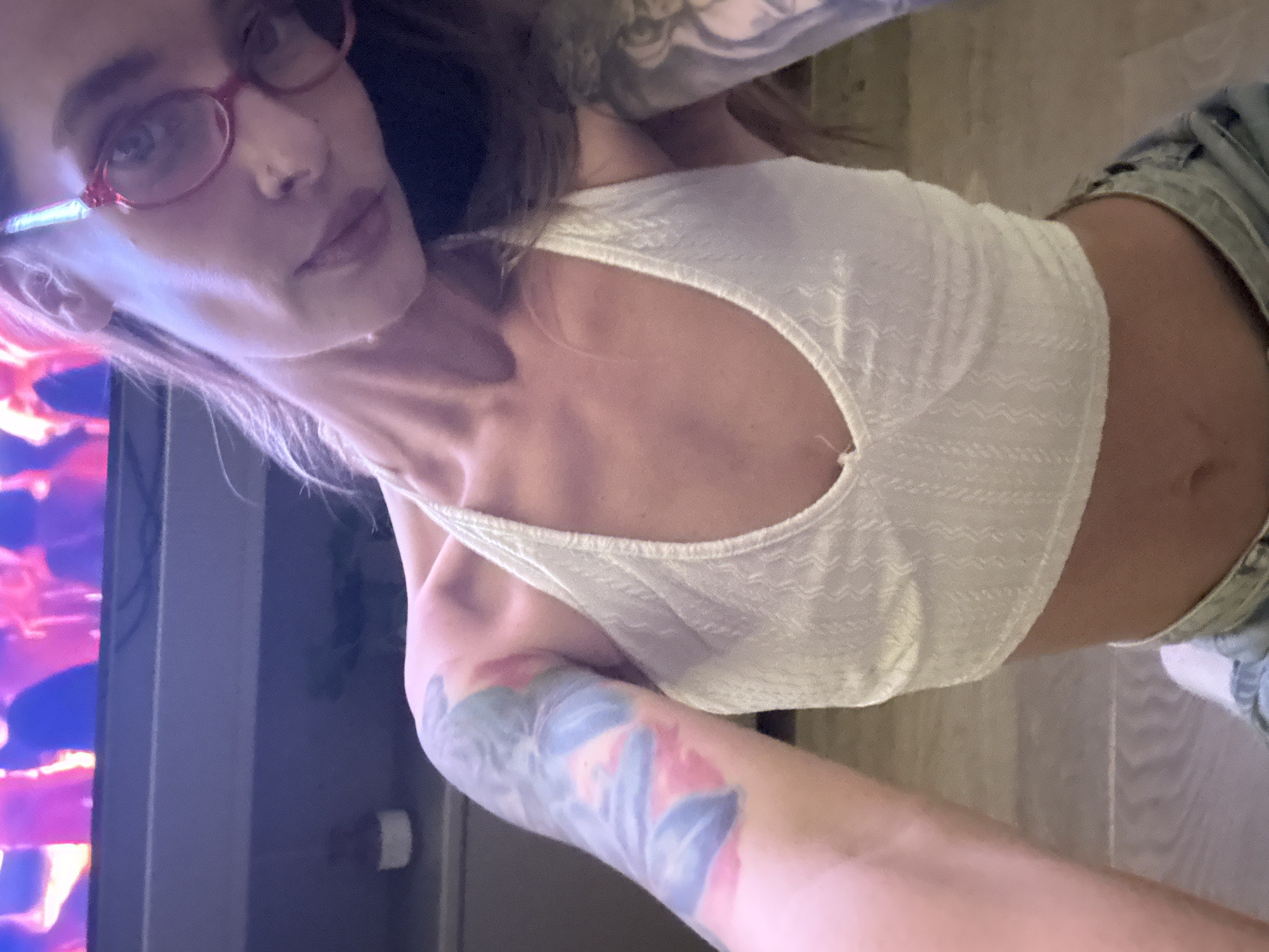 The image features a person with fair skin and light brown hair, wearing red-rimmed glasses, a white crop top, and denim shorts. The person has tattoos on their arms. To the left of the person, there is a television screen displaying a fire-like image. The background appears to be a room interior, with a light-colored floor. There is no specific information in the image to determine if the location is Tel-Aviv, Israel.