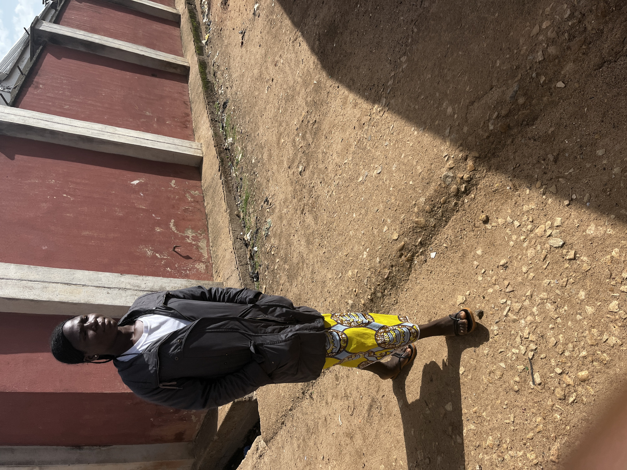 A young African person stands outdoors on a dirt path next to a red wall with concrete supports. The person is wearing a black jacket over a white t-shirt and brightly patterned yellow shorts with a blue and grey design. They are also wearing dark sandals. The person is looking off to their left, with their body angled slightly towards the camera. The scene appears to be in daylight, with harsh shadows cast on the ground, suggesting it is midday. The ground is rough and dry, with scattered small stones and some debris. The red wall is weathered and appears to be part of a building or structure. There is a concrete drainage channel or gutter running along the base of the wall. The overall setting is simple and utilitarian, possibly a residential area or a street in Jos, Nigeria. No text is visible in the image.
