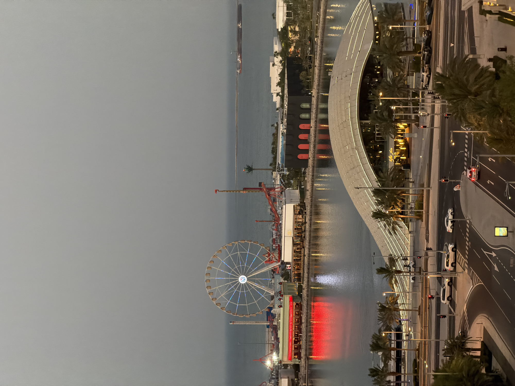 The image shows an aerial view of a waterfront entertainment district at dusk. In the background, a large Ferris wheel with illuminated spokes stands prominently against a hazy sky. Adjacent to the Ferris wheel are various amusement park rides, and waterfront buildings with glowing red and white lights. The water reflects the lights from the buildings. In the foreground, a modern, curved architectural structure with illuminated edges is visible, surrounded by palm trees. A road network with cars and traffic lights is also present in the foreground, with several vehicles in motion. The overall scene suggests a lively urban area with recreational facilities.