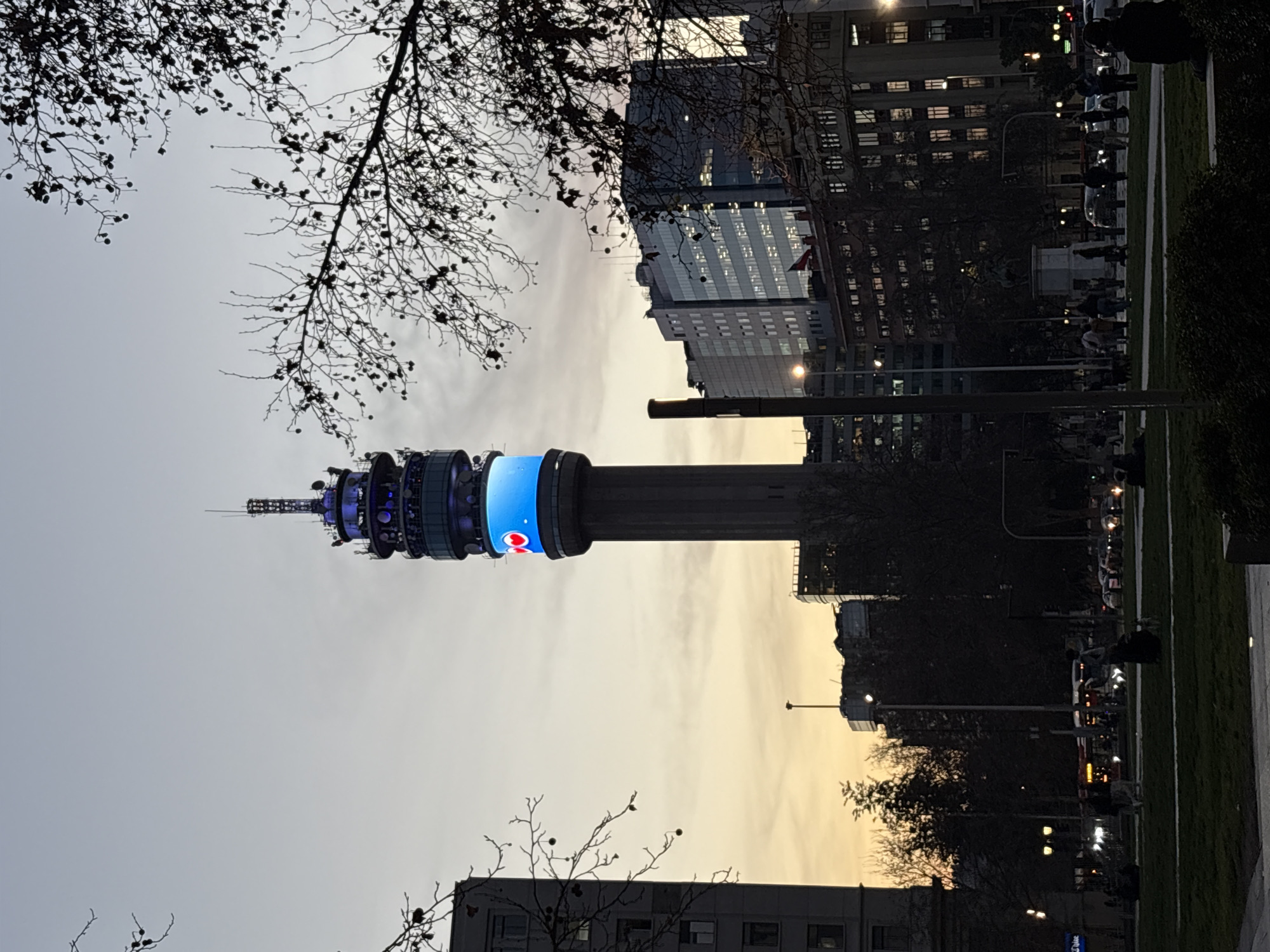 The image depicts the Torre Entel in Santiago, Chile, at dusk. The telecommunications tower is illuminated with blue lights and features a bright blue LED screen displaying two red hearts. The tower is centrally located, with a park in the foreground and several buildings visible in the background, including modern glass-fronted structures and older buildings. Silhouettes of trees frame the scene, their branches bare of leaves, suggesting a cooler season. In the park, several people are present, some walking dogs, and others sitting or standing. Traffic is visible on a street behind the park, with headlights and taillights indicating ongoing activity. The sky is a gradient of pale orange and grey, characteristic of twilight.