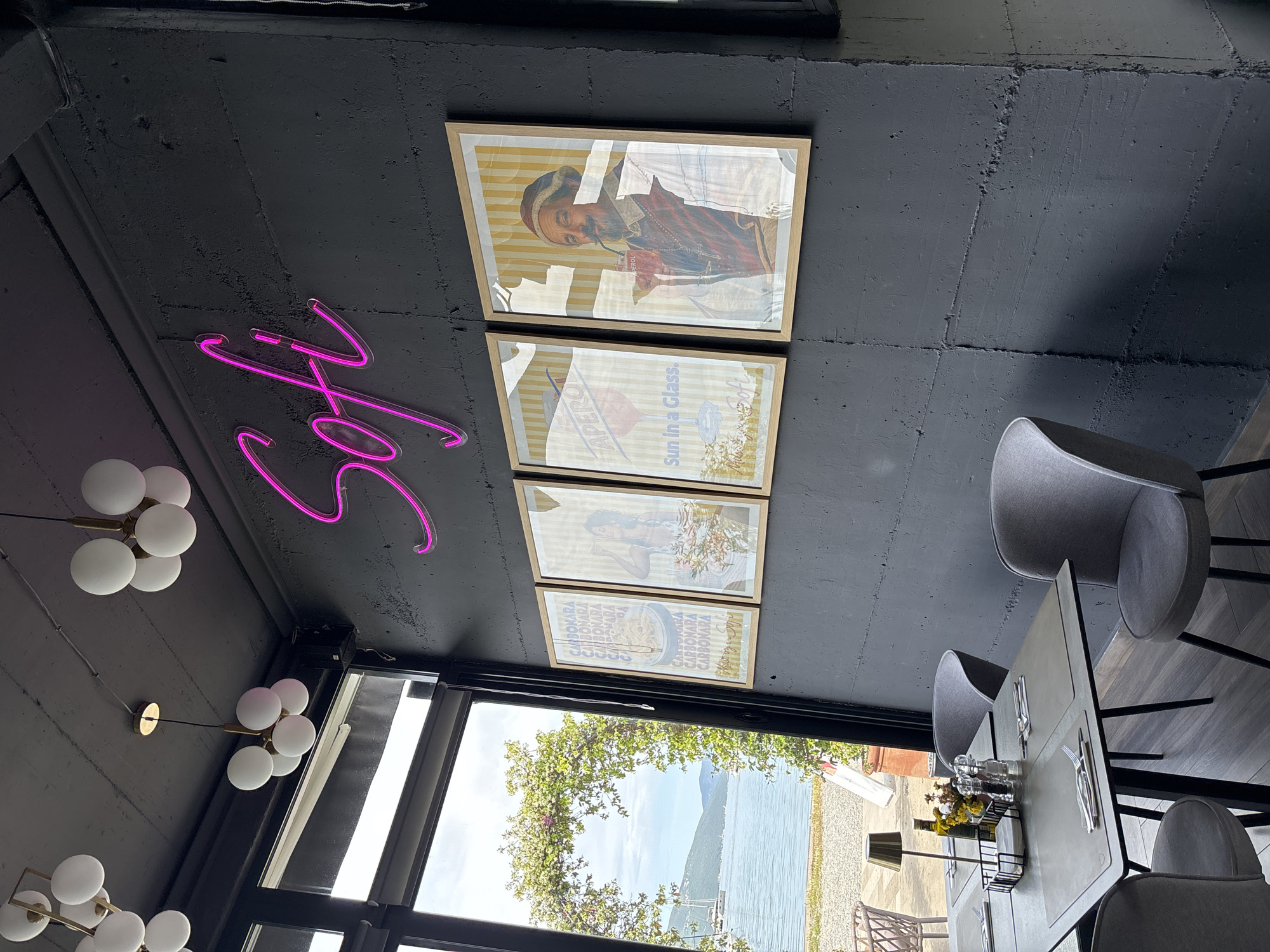 The image depicts an interior scene in Tivat, Montenegro. It captures a view of a modern restaurant or cafe, characterized by a dark, possibly concrete ceiling adorned with a pink neon sign spelling the name "Sofi." Below the sign, four framed art pieces hang aligned, with the upper-most containing a portrait of a man in historical clothing, while the lower three appear to feature vintage beverage or food advertisements.
To the left, a series of spherical, multi-bulb light fixtures hang down, illuminating the space. A large window offers a view of the Tivat Bay, framed by greenery, suggesting an outdoor seating area or a view of a waterfront. Inside, several sleek, modern gray chairs are arranged around a table set with cutlery, glassware, and a floral centerpiece. The table setting suggests a dining experience is in progress or prepared for guests.