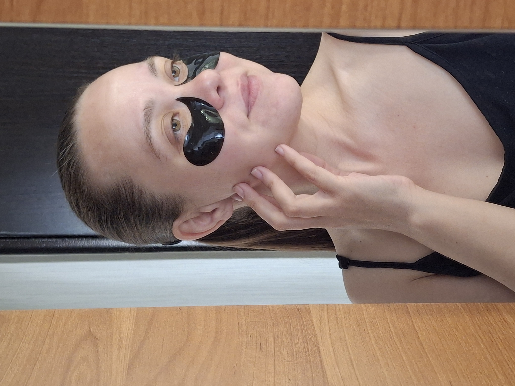 A woman is depicted wearing black eye patches while lying down on what appears to be a yoga mat. Her face is partially obscured by the eye patches, but her eyes are visible. She is touching her face with her right hand. The woman is wearing a black top with thin straps. The yoga mat is placed on a surface made of what seems to be a wood-like material. The location within the city of Chaikovsky, Russia, is not discernible from the visual information provided in the image.