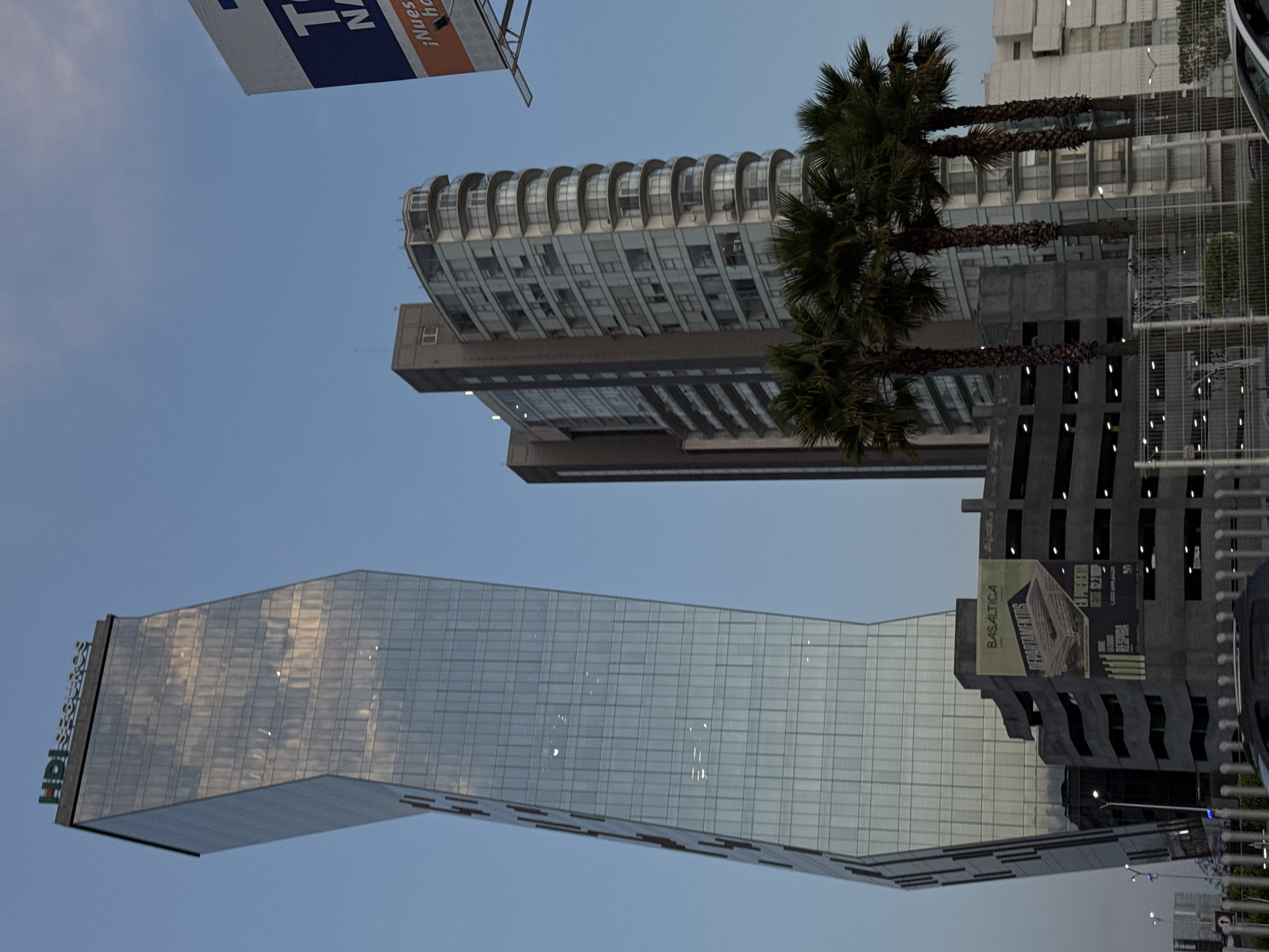 The image captures a view of modern architecture in the city of Puebla, Mexico. A sleek, glass-faced building with "HPI" branding dominates the left side of the frame, reflecting the sky. Adjacent to it is another building, possibly a parking structure, with a large advertisement banner. Tall palm trees stand to the right of these buildings, adding a touch of nature to the urban landscape. In the background, a residential building with a distinct curved roofline is visible. The sky is a clear, light blue.