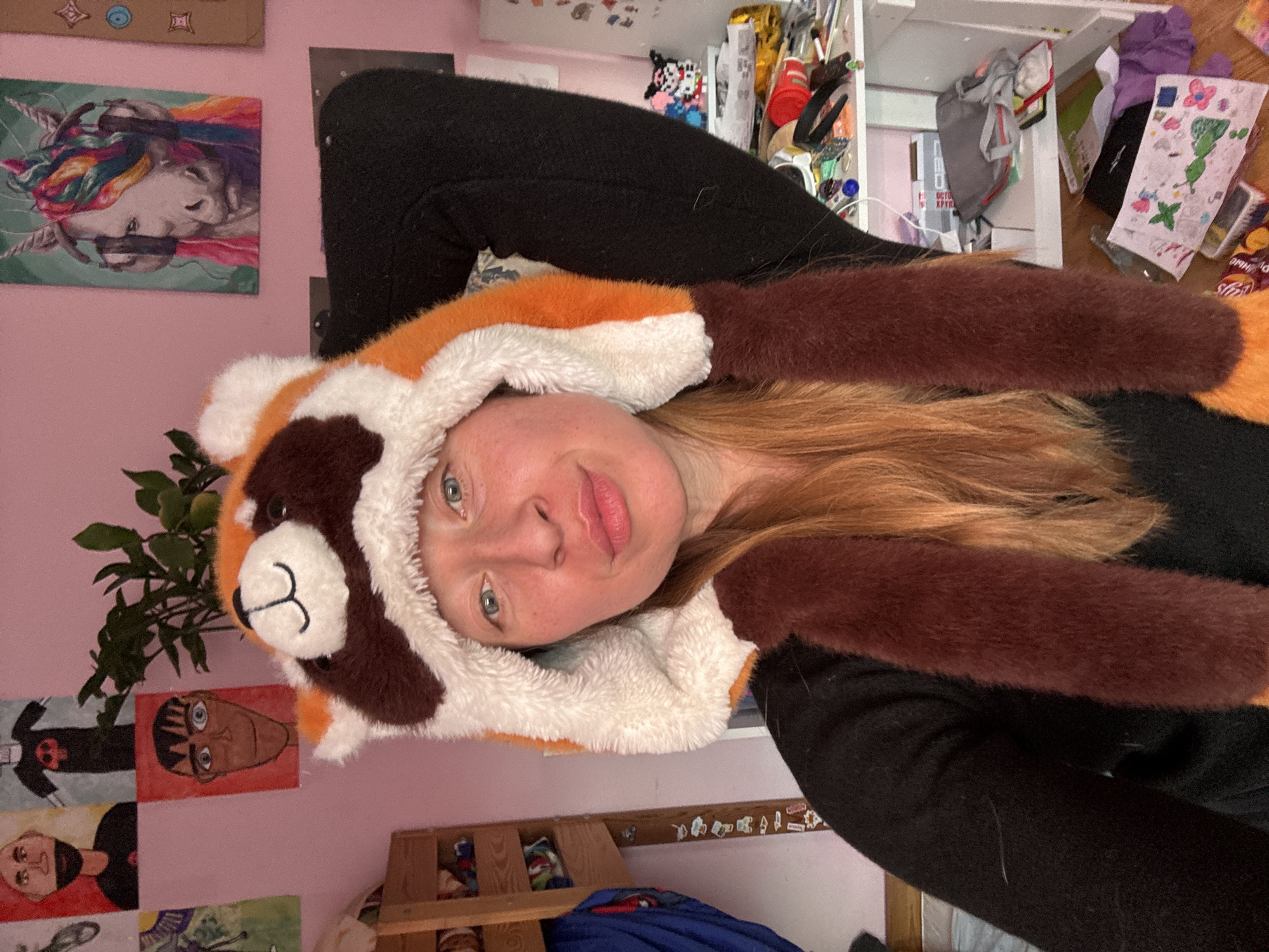 A woman wearing a red panda hat is indoors, presumably in a home. The background includes a pink wall with several framed pictures, a wooden shelf, and a shelf with miscellaneous items. No information is available to determine if the location is in Noviy, Russia.