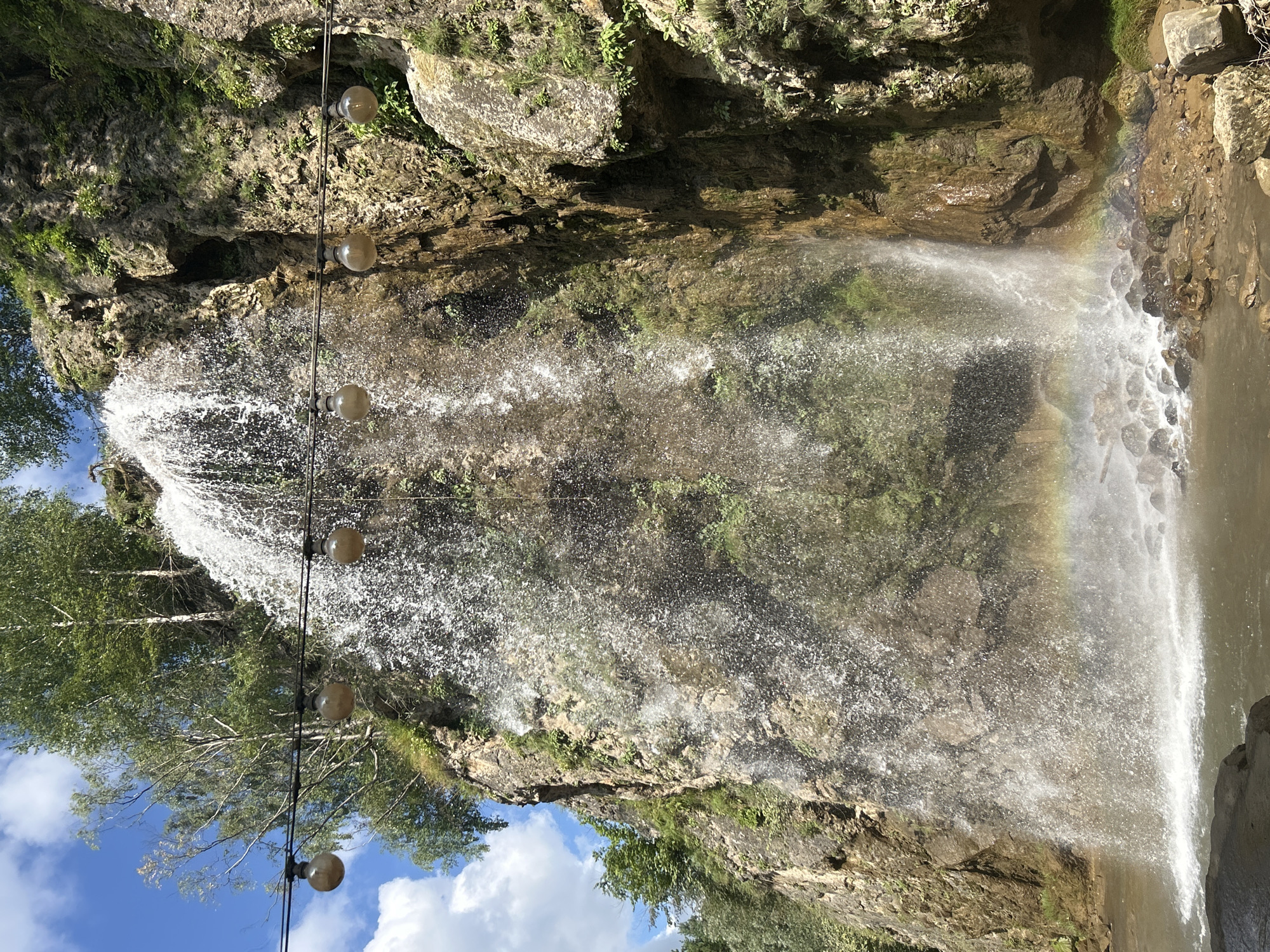 A wide, vertical shot captures a waterfall cascading down a rocky cliff face on a sunny day. The waterfall is the central focus, with water spraying outwards as it plunges into a pool below. A faint rainbow is visible in the mist at the base of the waterfall. Above the waterfall, several bare, spherical light bulbs hang suspended from a wire. The cliff is covered in patches of greenery and moss. The sky is blue with scattered white clouds. The scene appears to be a natural setting possibly within or near the city of Krasny Kurgan, Russia.