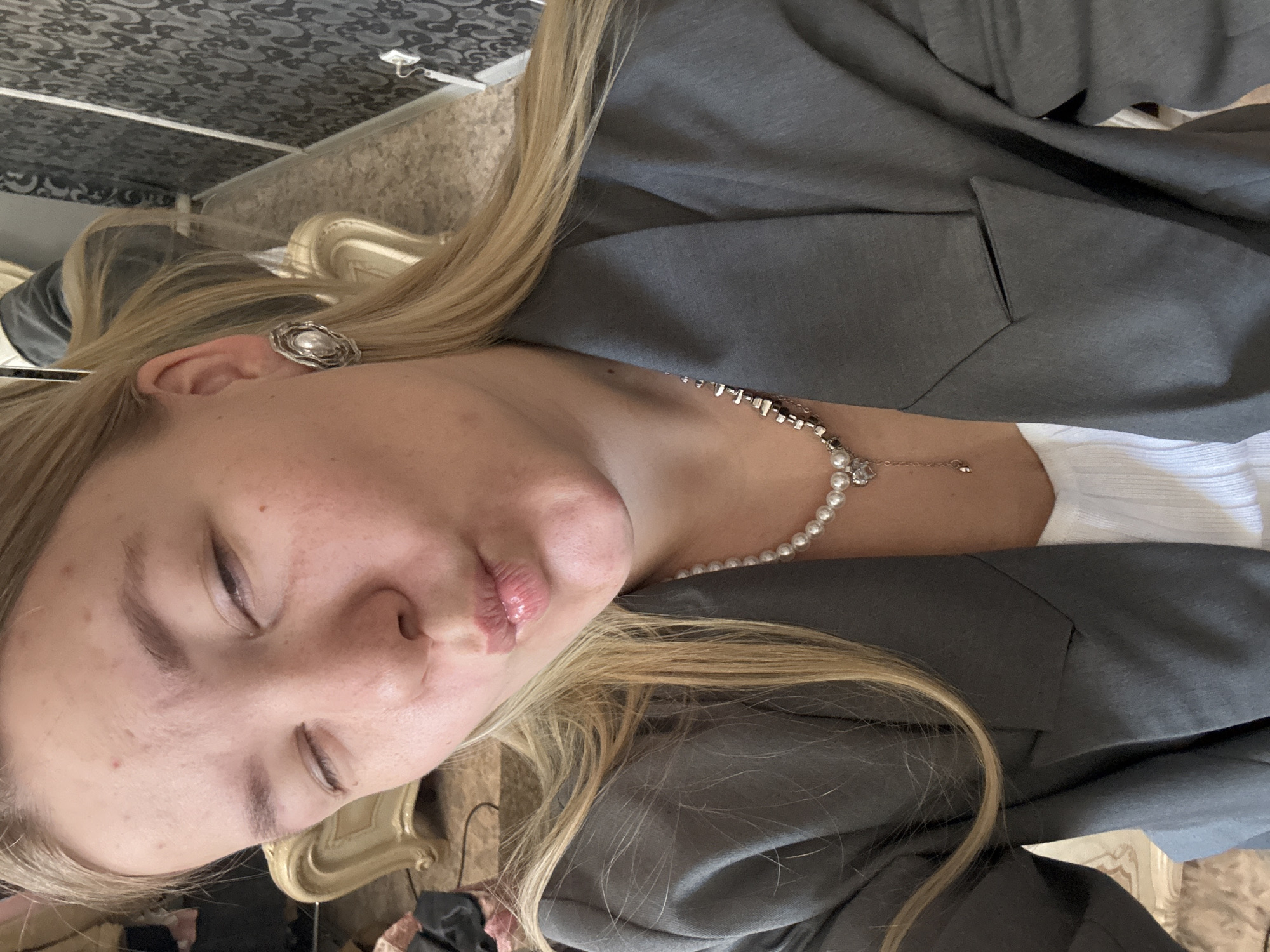 A young woman with closed eyes is wearing a gray blazer and a white top. She has blond hair and is wearing an earring and a pearl necklace with a diamond pendant. The background is a room with patterned wallpaper. It is impossible to determine if this image was taken in Chaikovsky, Russia.