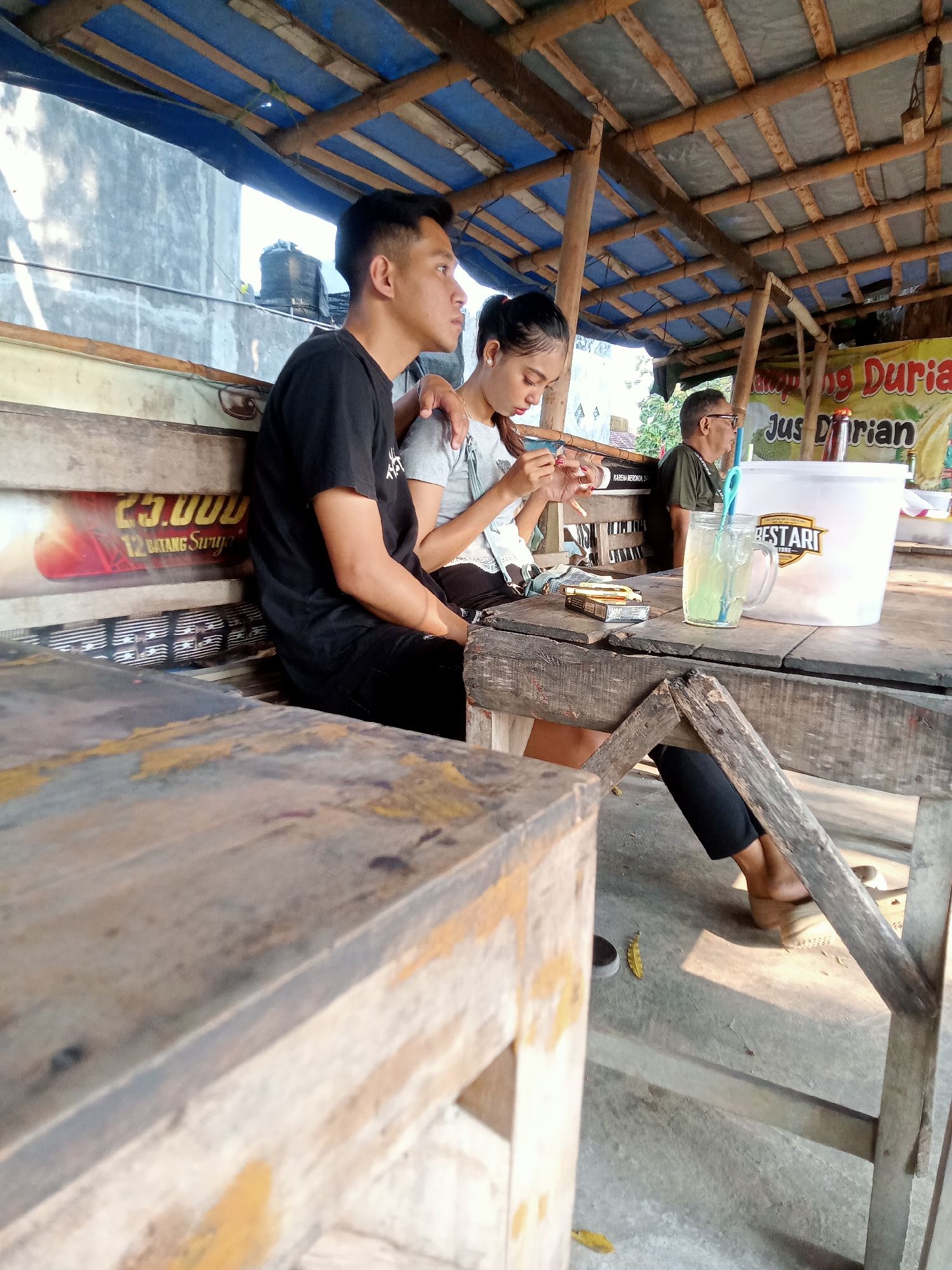 A young man in a black t-shirt and black pants is seated on a wooden bench. To his right, a young woman in a light gray t-shirt and dark pants is also seated. She holds a cigarette in her right hand. To the right of the woman, a man wearing glasses and a green t-shirt is seated. The three individuals are under a rustic wooden canopy with a blue tarp roof. A wooden table is in front of the woman and the man with glasses. On the table, there is a glass of light green liquid with a blue straw, a white bucket with a logo that reads "BESTARI STORE", and a pack of cigarettes and a lighter. In the background, there is a wall and foliage. A banner with text, including "Jus Durian", is visible in the background. The scene appears to be an outdoor seating area at a roadside stall, likely in Majan, Indonesia, given the presence of the durian juice and the overall aesthetic.