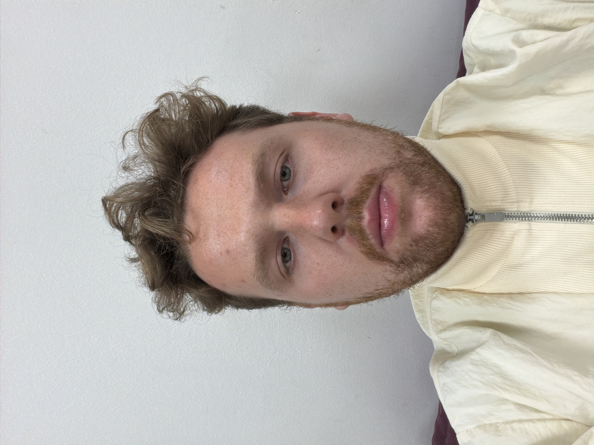 The scene depicts a close-up, head-and-shoulders portrait of one adult male subject. The subject has a light skin tone with visible freckles across the forehead and cheeks, greenish-blue eyes, and light brown or auburn curly hair that appears disheveled. He possesses auburn facial hair, including stubble or a short beard covering his chin and jawline. His lips are slightly parted and appear moisturized. The subject is wearing a cream or off-white collared jacket with a ribbed texture on the collar and front placket, secured by a silver-colored metallic zipper. The main body of the jacket has a smoother, slightly wrinkled texture. He is looking directly into the camera. The environment is an indoor setting with a plain, light-colored (white or off-white) wall serving as the background. A dark red or maroon surface, possibly upholstery from a sofa or chair, is partially visible behind his shoulders on both the left and right sides of the frame. There are no identifiable landmarks or features within the image to determine a specific location within the City of London, United Kingdom; it appears to be a private or non-descript interior space.