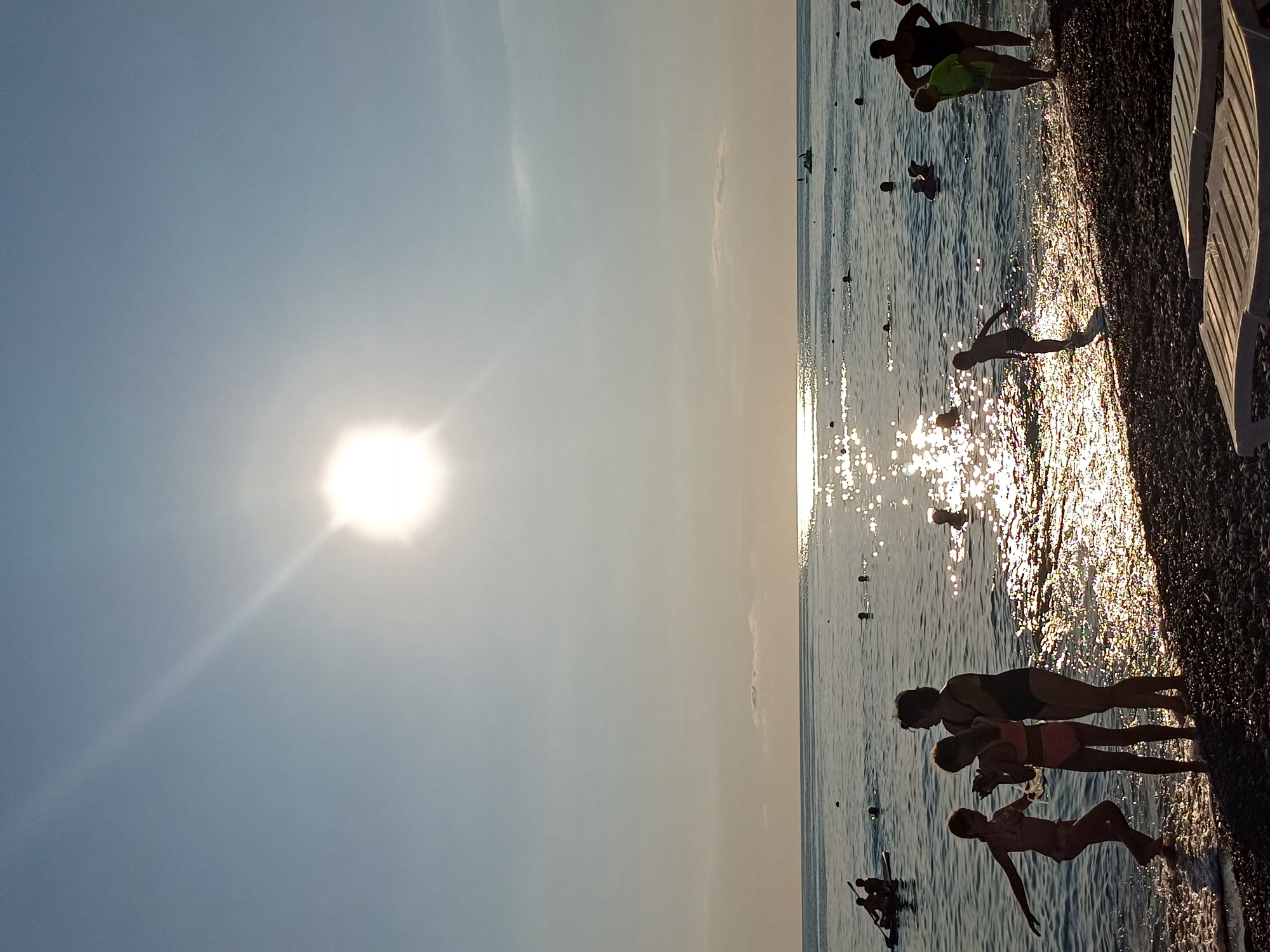 The image depicts a sunny day at the beach in Sochi, Russia. Silhouettes of several people are visible along the shoreline, with the sun setting in the background, casting a warm, golden glow on the water and the pebble beach.
In the foreground, three figures are gathered near the water's edge. Two appear to be adults, one in a dark swimsuit and another in a bright green top and shorts, possibly a parent and child. A third figure, a young child in a patterned swimsuit, is further down the beach, walking towards the water. In the middle ground, another silhouette of a person, possibly a child, is visible in the shallow water. Further out in the sea, more distant figures are scattered, some appearing to be swimming or wading. The sea is calm, with gentle waves lapping at the shore. The sky is a clear, pale blue, with a faint hazy effect from the bright sun.
The time of day is clearly late afternoon or early evening, given the low angle of the sun and the warm, extended shadows. The weather appears to be pleasant and warm, conducive to beach activities. The presence of beach chairs on the right side of the image suggests a resort or public beach area. The overall mood conveyed is one of relaxation and enjoyment of a summer day by the sea. There is no visible text in the image.